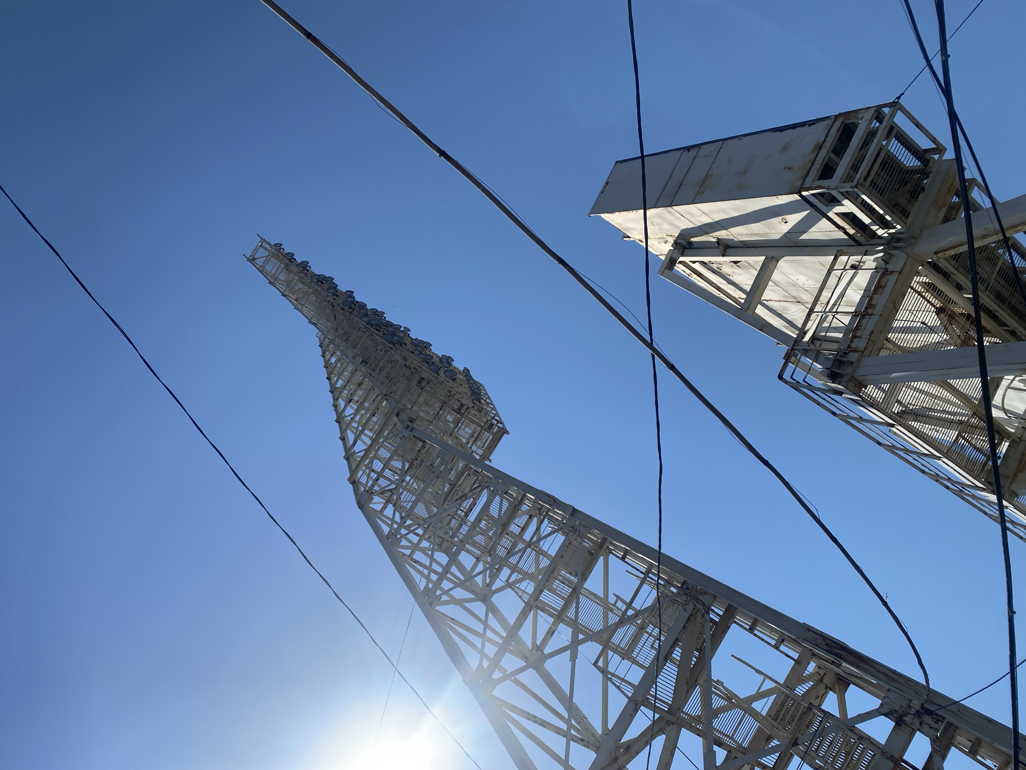 The image shows a low-angle view of two tall, white, lattice-structured towers against a clear blue sky. The tower on the left tapers to a point and has a series of lights or fixtures mounted at the top. The tower on the right is shorter and features a boxy enclosure at its peak. A network of overhead power lines is visible in the space between the towers and extending into the sky. The sun is positioned near the base of the left tower, creating a lens flare effect. Based on the provided information, this image may have been taken in or near the city of Заволжье, Russia.