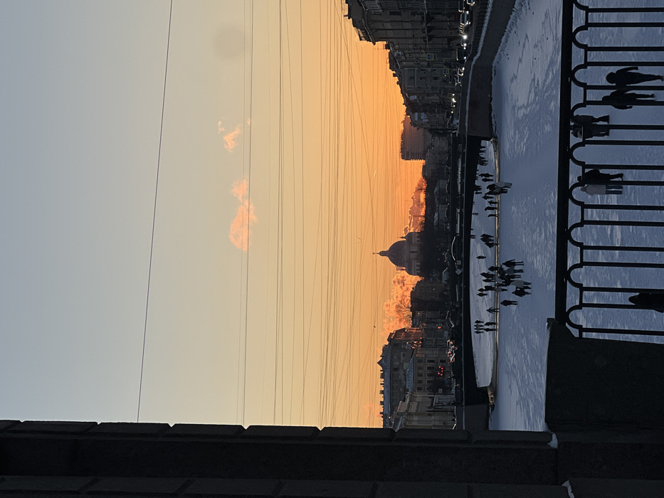 The image captures a wide-angle view of a frozen urban canal or river in Saint Petersburg, Russia, at sunset or sunrise during winter.
In the foreground, the lower portion of a dark, wrought-iron railing with vertical bars is visible, behind which several indistinct human figures are silhouetted against the lighter ice. To the immediate left, a large, dark vertical structure, possibly a stone building facade or bridge support, frames the scene.
The midground features a wide, flat expanse of a frozen waterway covered in snow and ice. Numerous people, appearing as dark silhouettes, are scattered across its surface, engaged in activities such as walking or skating. The waterway passes under at least one dark, arched bridge spanning its width. Along the banks, city buildings line the streets. On the right bank, a continuous row of multi-story buildings is visible, some with illuminated windows. Vehicles with headlights on are present on the street running parallel to the river.
The background is dominated by the silhouette of Saint Isaac's Cathedral, identifiable by its large, classical dome and cupola, situated centrally against the vibrant horizon. Other city buildings and structures extend along the horizon line.
The sky exhibits a clear gradient, transitioning from a light blue at the top to a prominent orange and yellow hue closer to the horizon, indicative of a low sun angle. Multiple thin, dark horizontal lines, consistent with utility wires or power lines, traverse the sky across the upper and middle sections of the frame. One small, indistinct cloud formation is visible in the upper-mid sky against the orange backdrop. The overall environment is cold, illuminated by natural low-angle light and artificial city lights.