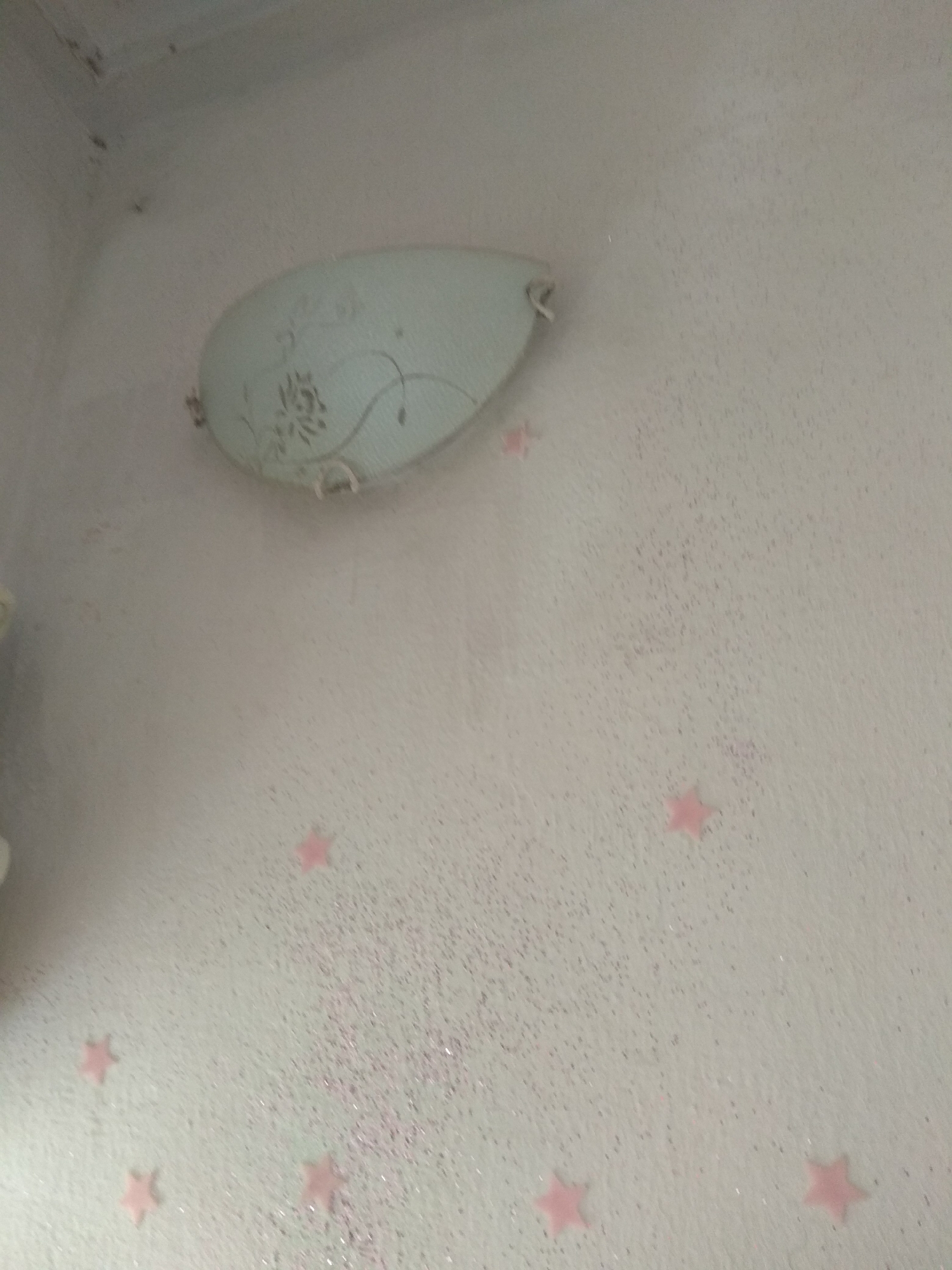 The image shows a close-up of a wall with a light fixture. The wall is painted a light color and appears to have a textured finish with small specks of glitter. There are several pink star-shaped decorations adhered to the wall. A light blue, oval-shaped light fixture with a floral pattern is mounted on the upper portion of the wall. The lighting suggests it might be daytime, as the light fixture appears to be off. The setting is likely an indoor room. No people are visible in the image, and no text is present.