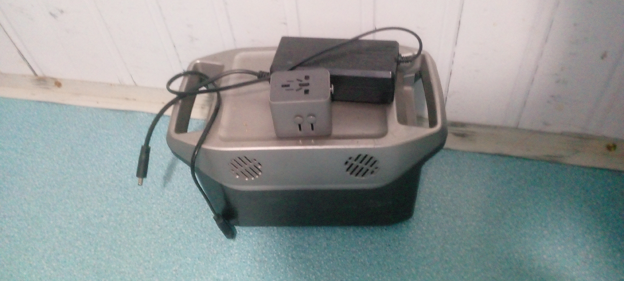 The image shows a portable power station with various adapters and cables plugged into it. The power station is gray and black, and it has ventilation grilles on the sides. A black power adapter is placed on top of the power station, and a gray universal travel adapter is plugged into the power station, with a cable connected to the power adapter. The power station is sitting on a light blue textured surface, and a white wooden wall is visible in the background. The lighting is dim, and the image quality is poor, suggesting it was taken indoors. There are no people or animals visible in the image, and no specific location in Indo, Nigeria, can be identified.