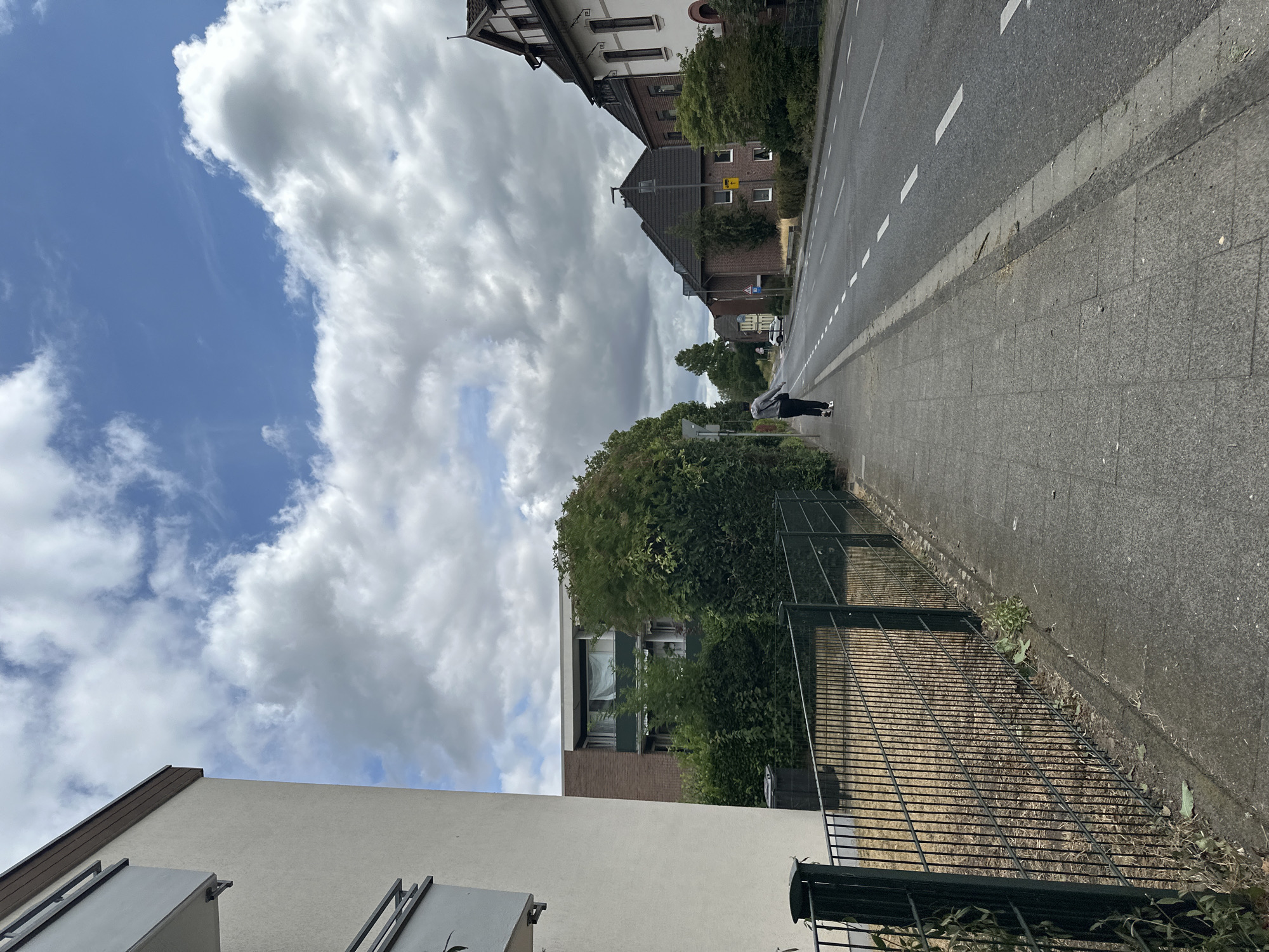 The image depicts a residential street scene in Krefeld, Germany, under a partly cloudy sky. In the foreground, a paved sidewalk runs alongside a dark metal fence and a textured building wall on the left. A lone individual, dressed in a grey hooded sweatshirt and dark pants, walks away from the camera on the sidewalk, heading down the street. The street itself is asphalt, marked with white dashed lines, and has a concrete curb. Adjacent to the street on the right are residential buildings with pitched roofs and multiple windows, interspersed with trees and hedges. The architecture suggests a suburban or older urban neighborhood. The overall impression is of a typical weekday scene in a German town.