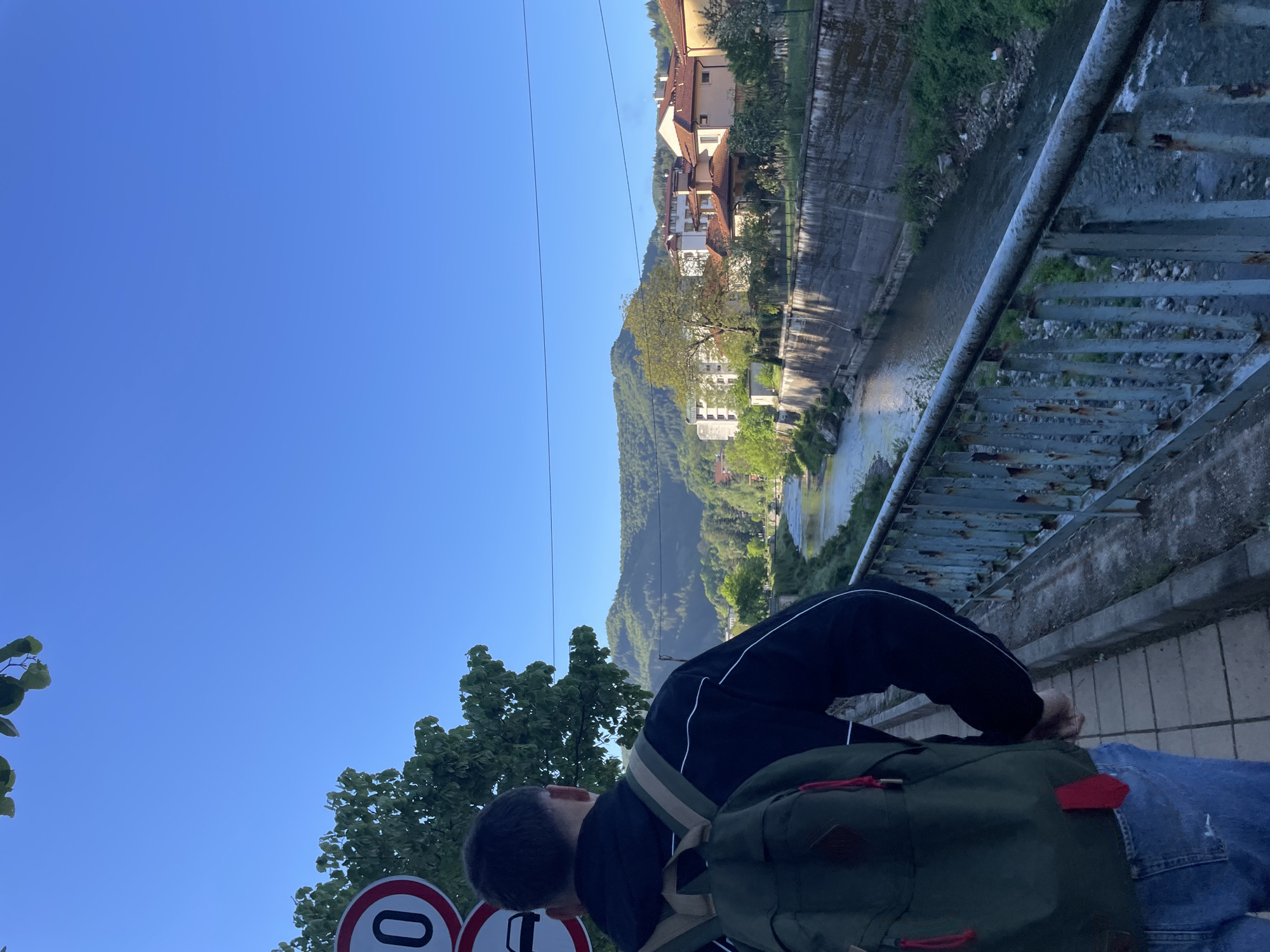 A medium outdoor shot shows a person wearing a black jacket with white trim, blue jeans, and a dark green backpack leaning over a bridge railing. The person appears to be looking down at a river running alongside a concrete wall and buildings. The location is Zlatograd, Bulgaria. A road sign is visible in the foreground. The sky is clear and blue.