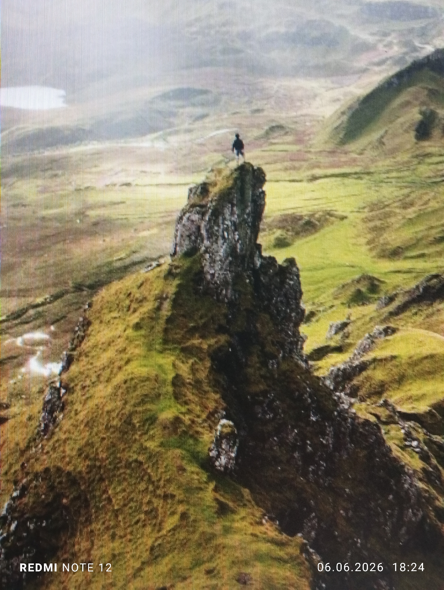 The image displays a rugged, expansive landscape, geographically identified as being within the city of Burgas, Bulgaria. A lone human figure, appearing as a dark silhouette, stands prominently atop a tall, craggy rock pinnacle situated in the mid-ground. The individual faces away from the viewer, observing the distant terrain. The environment is characterized by undulating hills and valleys, predominantly covered in patches of green and brownish vegetation interspersed with visible rocky outcrops. A calm body of water is observable in the upper left background, reflecting the ambient light. The sky is overcast or hazy, diffusing the light. The lower left corner contains a digital watermark reading "REDMI NOTE 12", and the lower right corner displays "06.06.2026 18:24", indicating a date and time. Vertical scan lines are visible across the entire image, suggesting a photograph of a screen or a similar digital artifact.