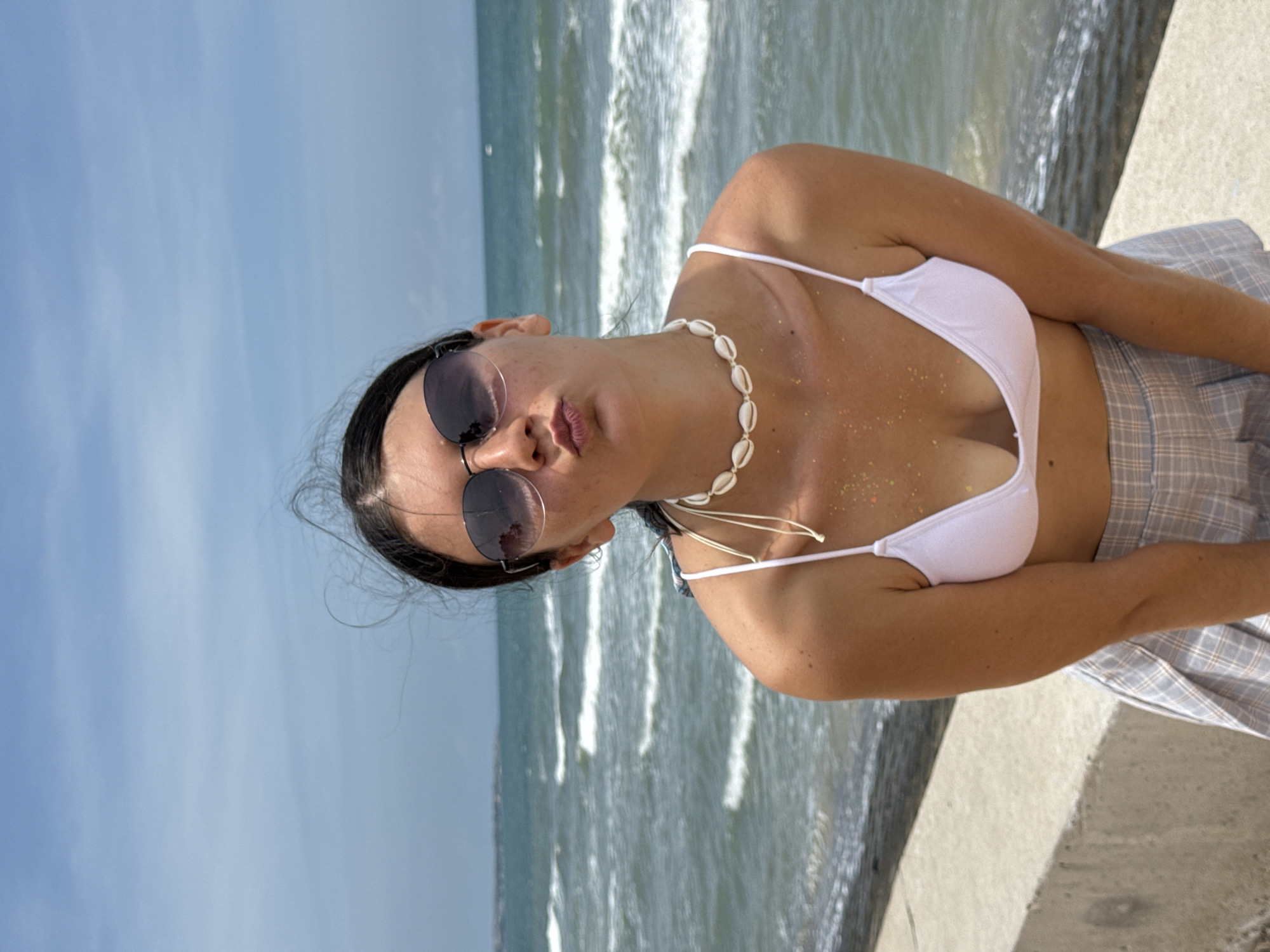 A woman with dark hair, wearing sunglasses, a white bikini top, and a plaid skirt stands on a concrete ledge facing the ocean. She is puckering her lips and has a shell necklace on. There are waves and a blue sky with some clouds in the background. The location is Phu Thuy Ward, Vietnam.