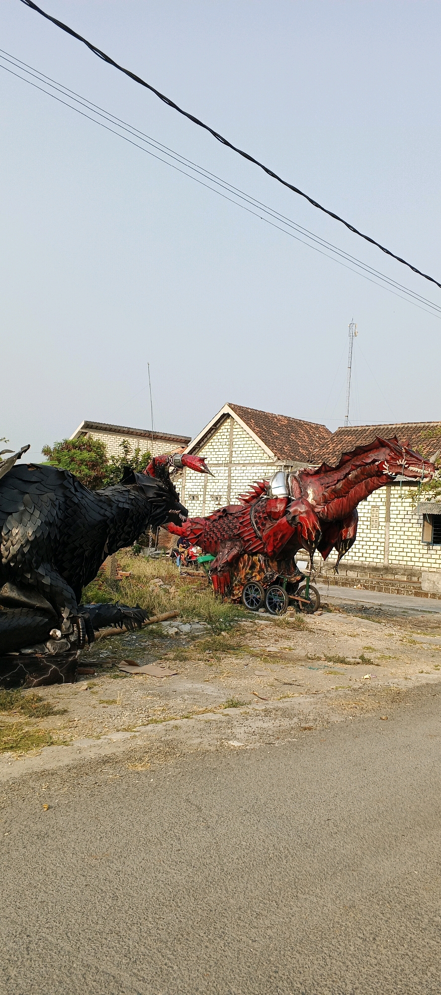 The image depicts two large, dragon-like sculptures made of what appears to be repurposed materials like tires and metal. One sculpture is black, resembling a wolf or a similar creature with sharp teeth and claws, positioned on the left. The other sculpture is red, detailed with scales and spiky protrusions, appearing more like a traditional dragon. This red sculpture is mounted on a wheeled cart, suggesting it might be mobile. The scene is set outdoors, with a dusty road in the foreground and grassy, unkempt terrain behind the sculptures. In the background, there are buildings constructed with white bricks and red tile roofs, indicative of a rural or suburban Indonesian setting. Electrical wires are visible overhead against a clear, pale sky. The general atmosphere suggests a display or perhaps a festive event. The location is identified as Latukan, Indonesia.