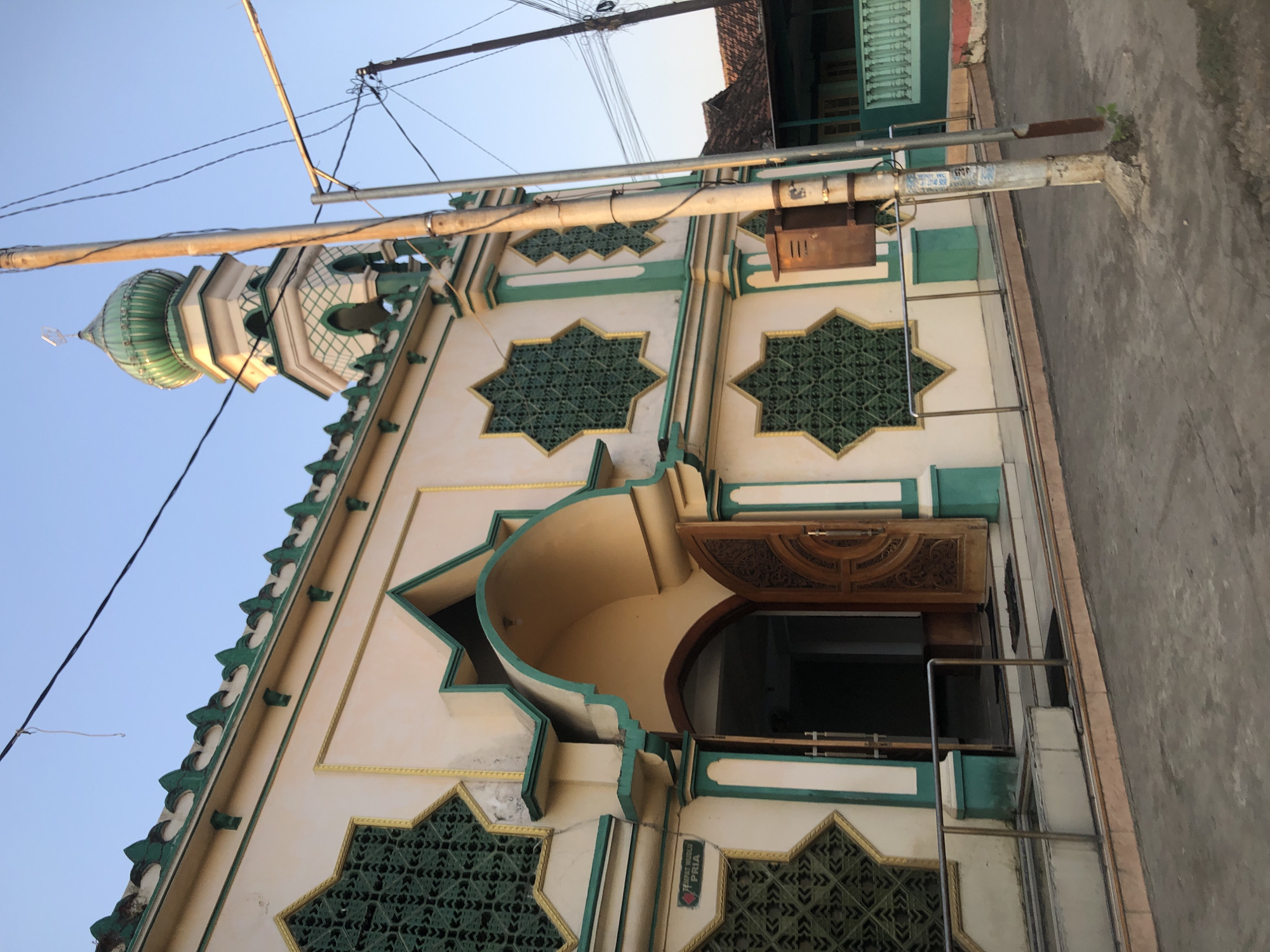 The image shows the exterior of a mosque with a green dome and minaret, located in the city of Keboguyang, Indonesia. The mosque has a white facade with green and gold accents, featuring star-shaped windows with intricate green lattice work. An arched entrance with a wooden door is visible on the right side of the building. A sign above the entrance indicates "TEMPAT WUDLU PRIA," meaning "Men's Ablution Area." The mosque is situated next to a street, with utility poles and wires in the foreground. In the background, a traditional Indonesian house with a tiled roof can be seen. The sky is clear and blue, suggesting it is daytime.
