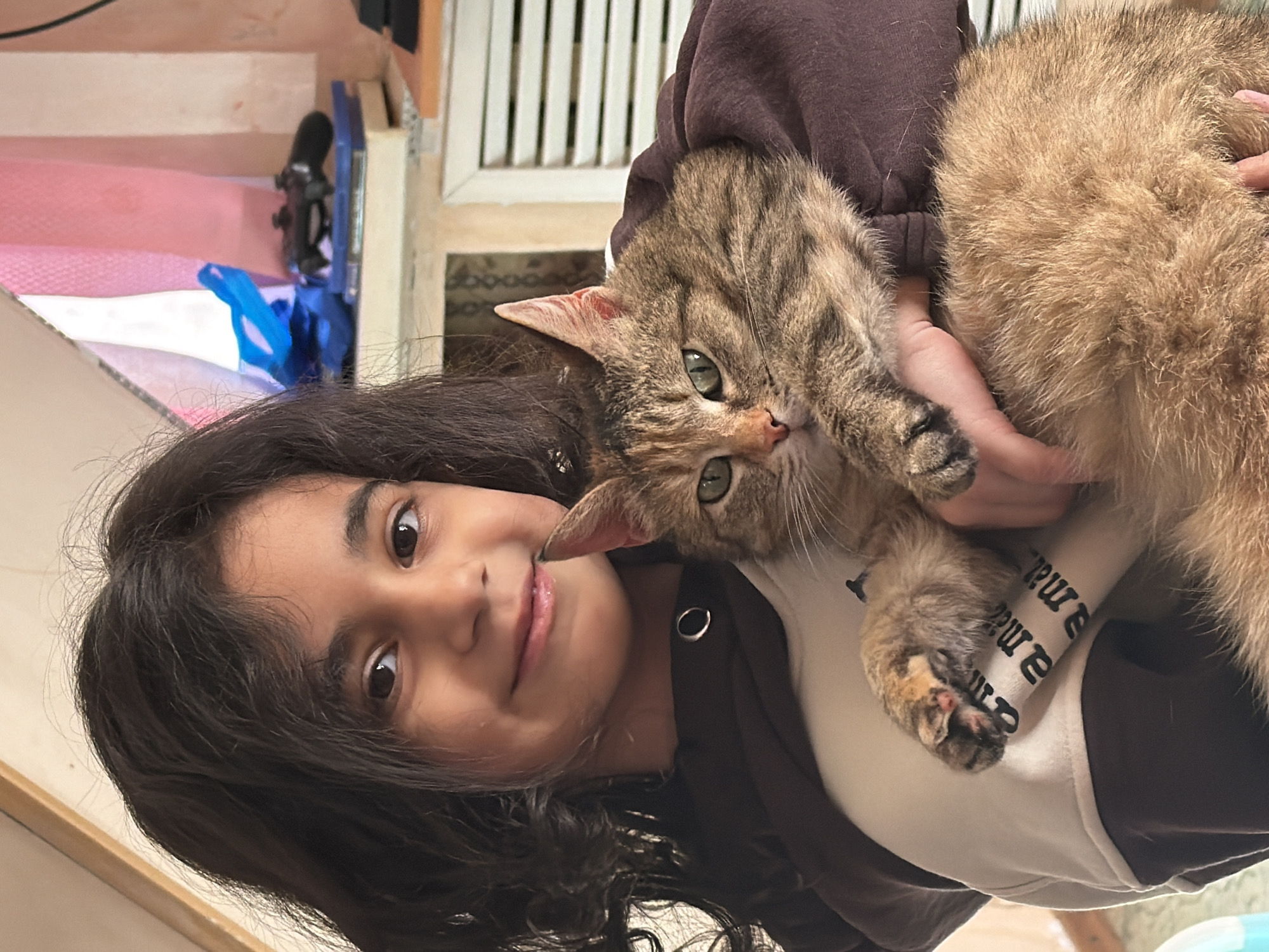 A young girl with long, dark, wavy hair is pictured indoors, holding a tabby cat with green eyes. The image is rotated 90 degrees clockwise, suggesting a selfie orientation. The girl is looking towards the camera with a gentle smile, her dark eyes open wide. She appears to be of elementary school age, with a warm skin tone. She is wearing a dark brown hooded top over a lighter shirt.
The cat, with mottled brown, black, and ginger fur, is nestled comfortably in the girl's arms, its head tilted towards the camera. Its left eye is clearly visible, and its right eye is partially obscured by its nose. The cat's paws are visible, one resting on the girl's shirt.
In the blurred background, there are hints of an indoor setting, possibly stairs with pink carpeting or a fabric runner, and some indeterminate objects including what looks like a black microphone or camera on a stand, and a blue item. A white slatted structure, possibly a cabinet door or room divider, is also visible. The lighting suggests daytime.
On the girl's light-colored shirt, partial text is visible, appearing to spell out "a ma" and "mama" multiple times in a repetitive pattern, written in black serif font. The scene conveys a moment of affection and companionship between the girl and her pet in an indoor setting in Таскино, Russia.