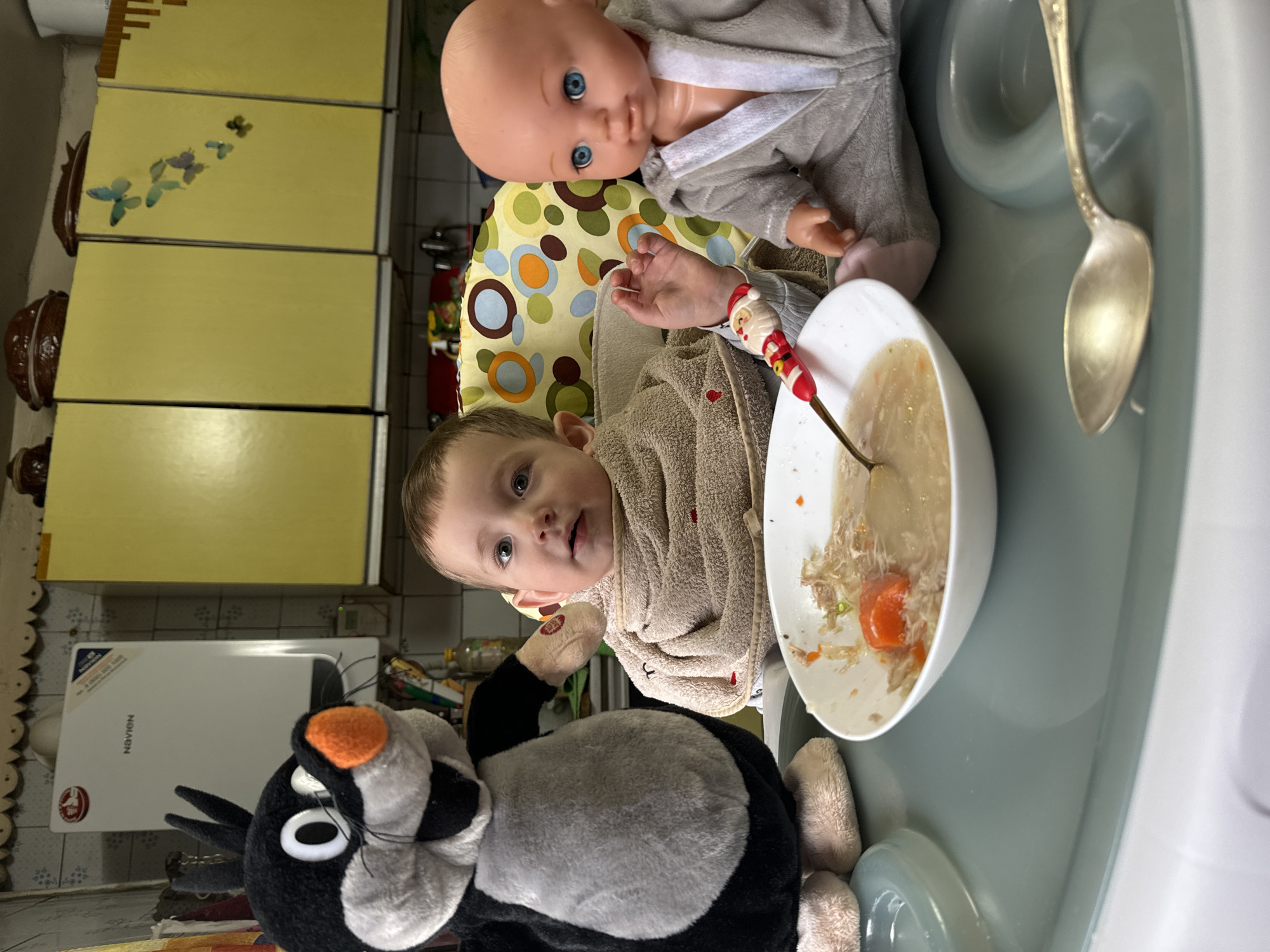 A baby sits in a high chair, facing the camera. The baby has light brown hair and light skin. The baby is wearing a beige bib. A black and white stuffed Tweety Bird is to the left of the baby. A bowl with soup is in front of the baby, and a spoon with a Santa Claus figure is in the soup. Another silver spoon is to the right of the bowl. A doll with blue eyes is sitting behind the baby. The high chair has a yellow and green patterned cushion. The background includes yellow cabinets, and a white Navien water heater. The location is Unknown, Unknown.