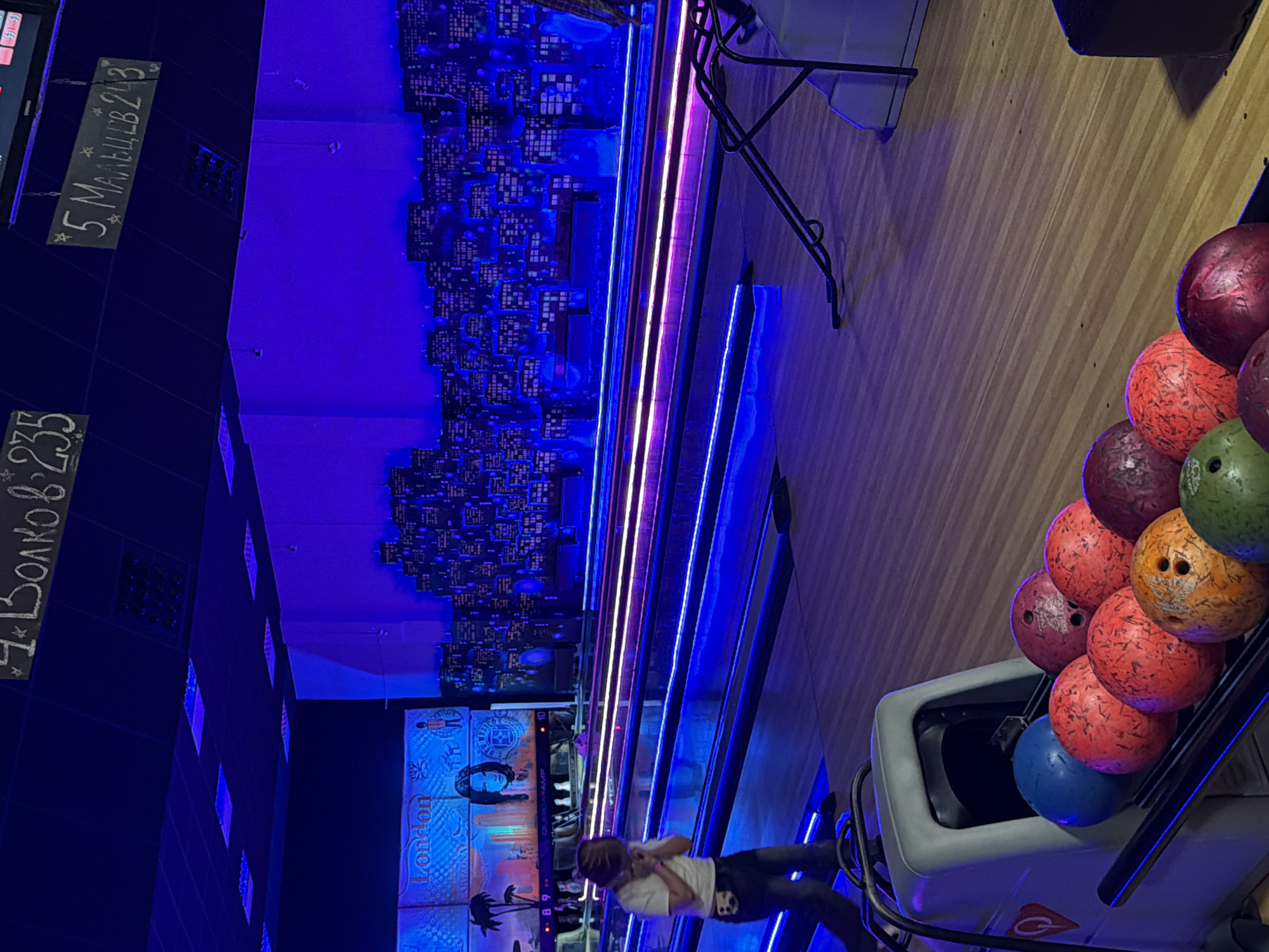 This image depicts an active bowling alley in Izhevsk, Russia, characterized by its vibrant, atmospheric lighting. The scene is illuminated primarily by blue and purple neon lights, suggesting an evening or nighttime setting.
In the foreground, a person, possibly a child or young adult, is standing near the bowling ball return area, wearing a white t-shirt and dark pants. A rack of various bowling balls in shades of red, orange, green, blue, and purple is prominently featured, indicating an ongoing game or preparations for one. The bowling lanes extend into the background, flanked by parallel strips of neon lighting.
The walls are decorated with a striking blue-lit cityscape mural, resembling a stylized skyline. Below this, there are decorative panels that feature text such as "London" and "Arizona," contributing to a global or themed aesthetic. Hanging above the lanes are two chalkboard-style signs displaying scores and names in Russian. One sign reads "4 BoлKoв 235" (4 Volkov 235), and the other "5 МАЛЬЦЕВ 243" (5 Maltsev 243), indicating high scores from participants with Russian surnames. The overall ambiance is lively and recreational, typical of a modern bowling center.