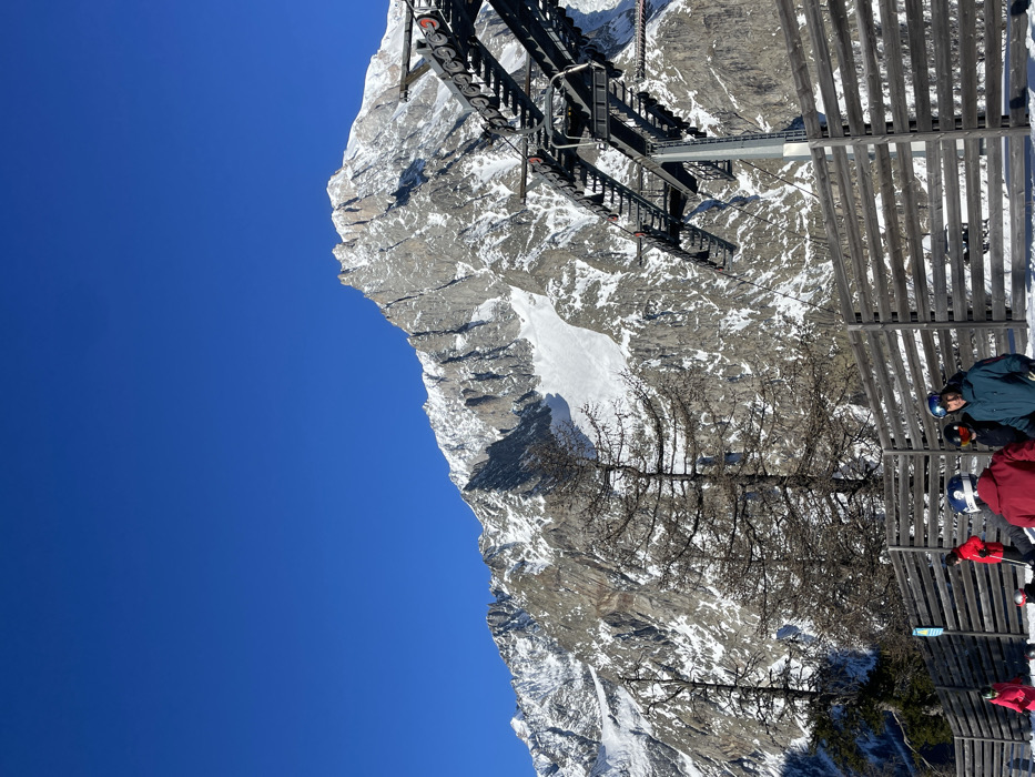 This image depicts a sunny winter day in a mountainous ski resort in Courmayeur, Italy. The scene is dominated by a vast, clear blue sky on the left, contrasting with rugged, snow-covered mountains that fill the right and central parts of the frame.
In the foreground, on the right side, several people dressed in winter jackets and helmets are visible. They appear to be standing or waiting near a tall wooden fence or barrier, suggesting they are at a ski lift station or a viewing area. Above them and extending across the upper-mid section of the image is a large, dark structure of a ski lift, possibly a gondola or chairlift mechanism, with its operational components clearly visible.
The mid-ground features a steep, rocky mountain slope partially covered in snow. Sparse, bare-branched trees, typical of an alpine winter landscape, are scattered across this section, adding texture. A prominent patch of smooth, white snow stands out against the darker rocks of the mountain.
The background reveals more snow-dusted mountain peaks under the bright, cloudless sky. The crisp light indicates a clear day, likely in the morning or early afternoon, with perfect conditions for winter sports. The overall atmosphere is one of serene natural beauty combined with the activity of a ski resort. No discernible text is visible.