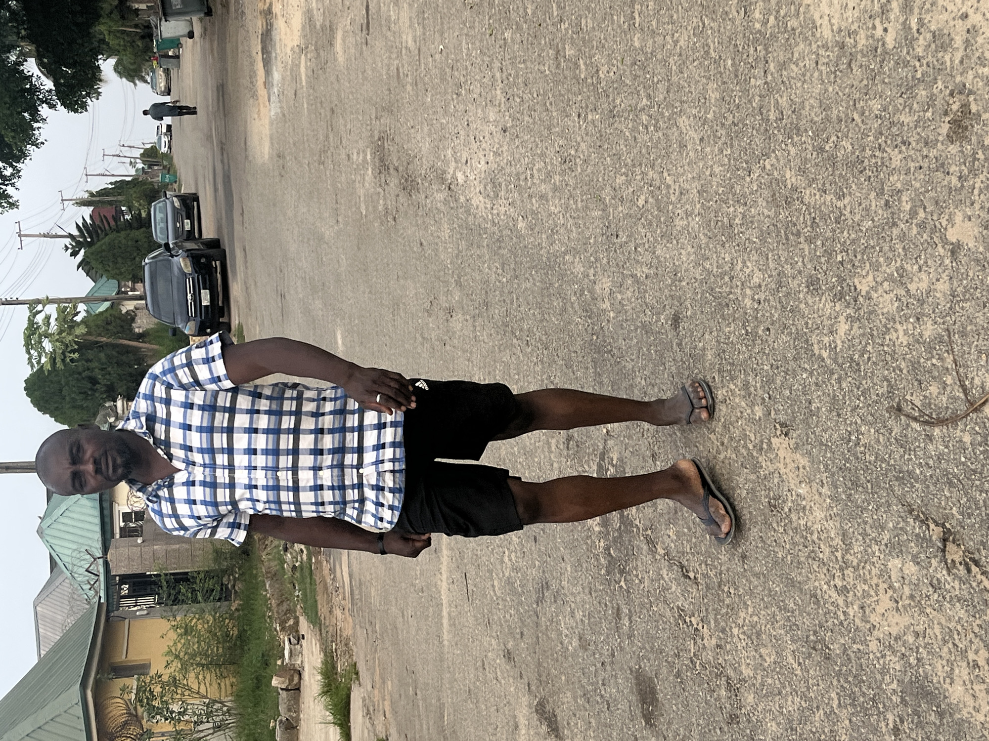 The image displays a man standing on a paved road in what appears to be a residential or semi-urban setting in Abuja, Nigeria. The primary subject, a man with dark skin, a shaved head, and a beard, is positioned in the foreground. He is wearing a blue and white plaid short-sleeved button-up shirt, black shorts with an adidas logo visible on the left leg, and dark flip-flops. He stands with his left hand on his hip and his right arm relaxed, looking directly at the camera with a neutral expression.
The road beneath him is textured and shows signs of wear, with visible cracks and aggregate. In the background, the road stretches further, flanked by houses, trees, and utility poles with overhead wires. Several dark-colored SUVs are parked along the road. Another person, dressed in dark attire, can be seen walking away from the camera in the far background. The buildings feature various architectural styles, including houses with green roofs and fenced perimeters, one with barbed wire.
The scene occurs during daytime under an overcast or hazy sky, resulting in soft, diffused lighting and an absence of sharp shadows. The overall atmosphere suggests a routine street environment. Aside from the man posing and the distant walker, no other significant activity is evident. Vehicle license plates are present but not legible.