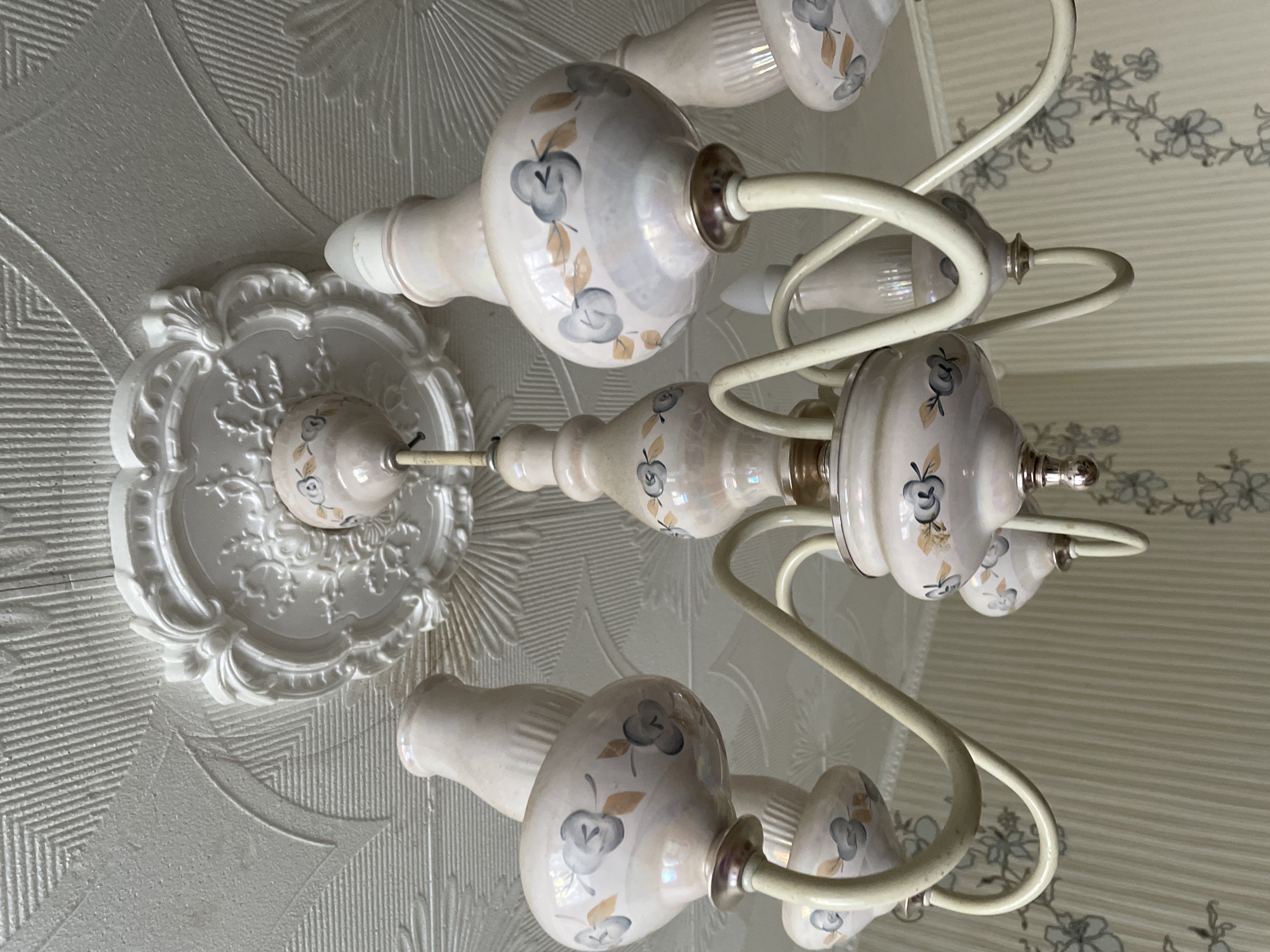 A vintage, cream-colored chandelier with floral motifs hangs from a textured white ceiling. The chandelier features multiple arms, each supporting a decorative light fixture made of ceramic. The ceramic shades are bulbous with a pearlescent finish and adorned with hand-painted blue and tan floral designs. The ceiling has a decorative white rosette medallion where the chandelier is attached. The wallpaper in the background is striped with a floral pattern. The scene is indoors, likely in a residential setting, with no people or animals present.