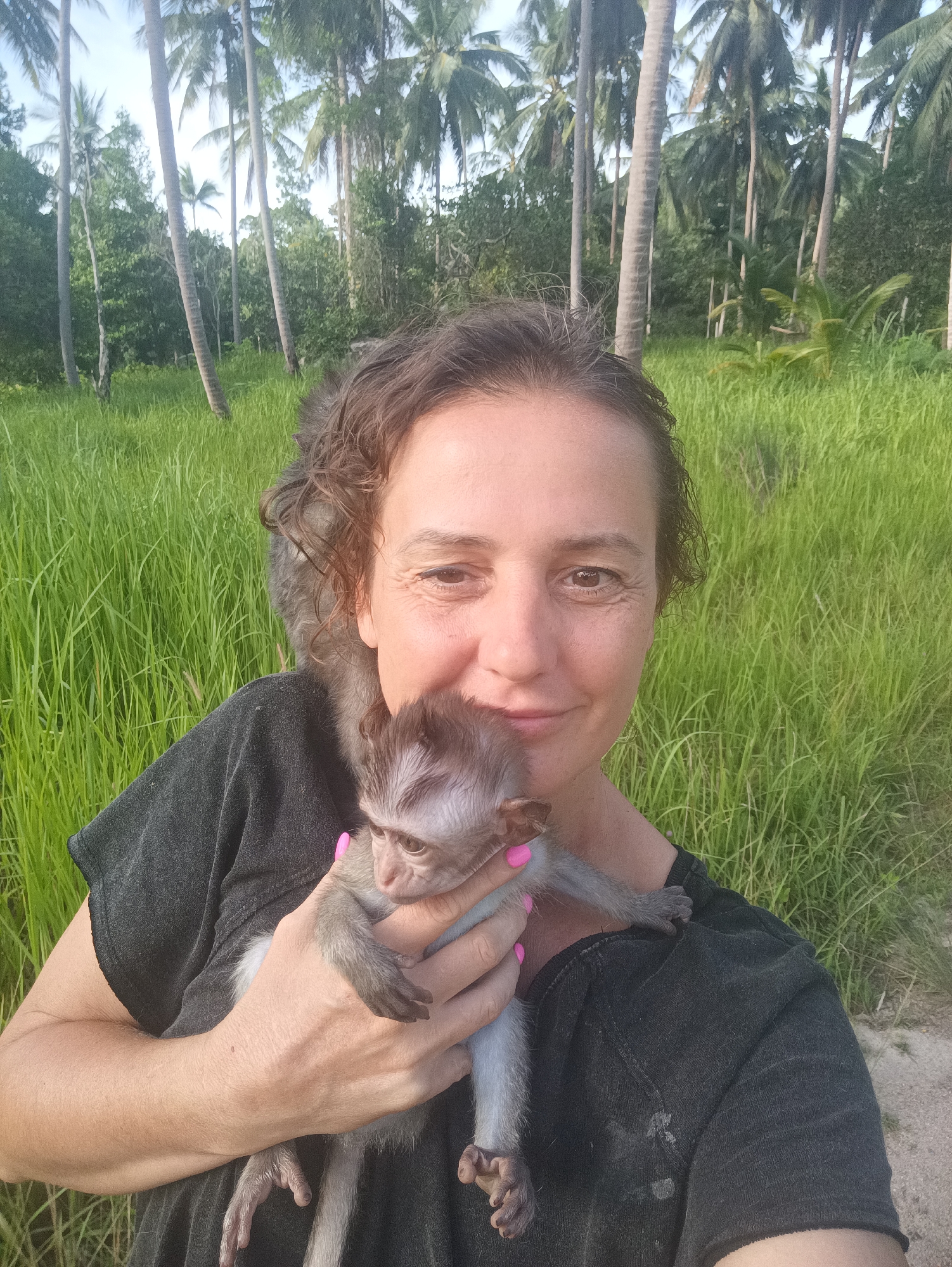A light-skinned woman poses outdoors, holding a monkey in her arms while another rests on her shoulder. The woman has short, curly, dark brown hair and is wearing a dark gray shirt. Her fingernails are painted bright pink. The monkey in her arms is small and gray with dark eyes and a striped pattern on its head. The monkey on her shoulder is facing away from the camera. The background consists of tall, green grass and a grove of palm trees. The location is Unknown, Unknown.