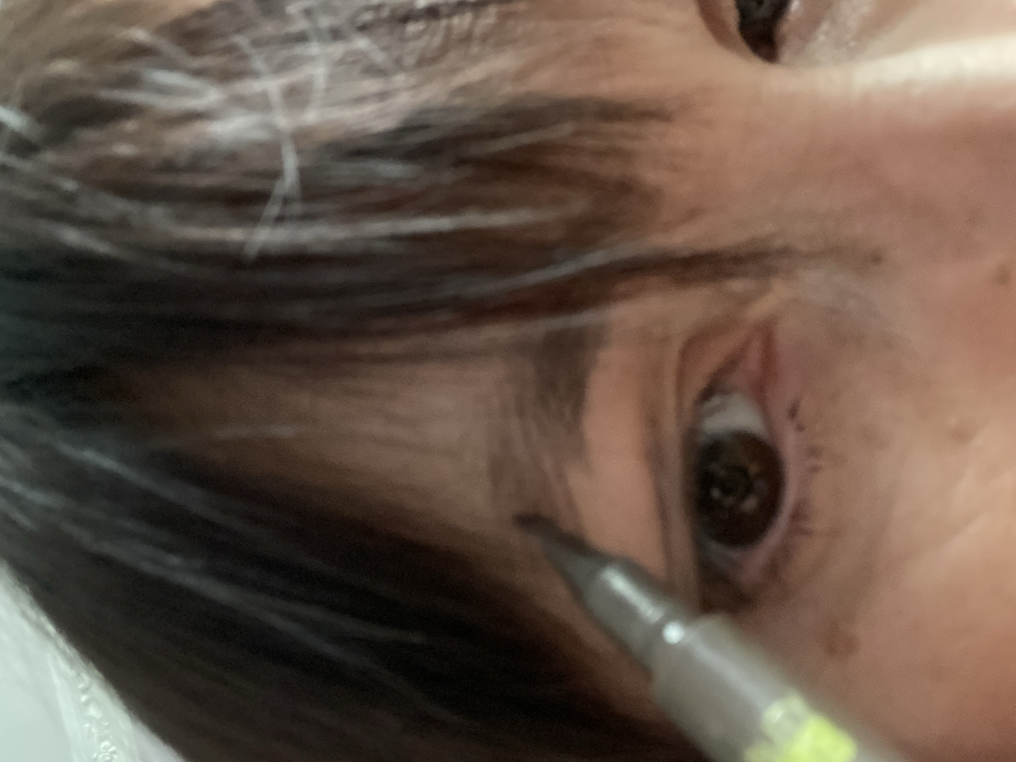 The image is a close-up of a person's eye area, focusing on the left eye and eyebrow. A cosmetic pencil is being held near the eyebrow, suggesting the person is applying makeup. The individual has dark hair that partially covers their forehead and some freckles are visible on the skin. The background is blurred, but appears to be indoors with artificial lighting. There is no information to suggest this image is located in Novokuznetsk, Russia.