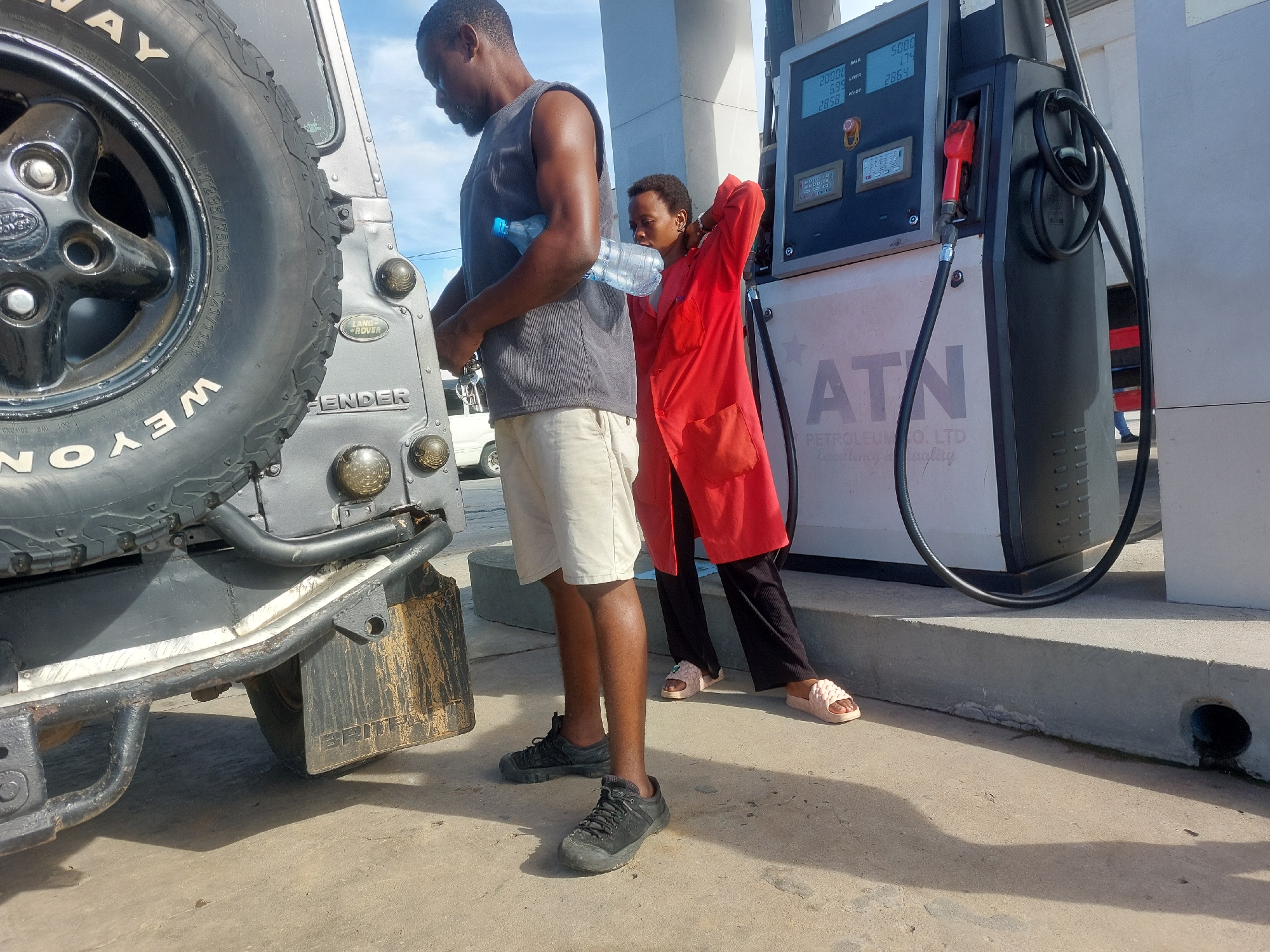 The image captures a scene at a gas station in Dar es-Salaam, Tanzania. A Land Rover Defender is positioned at a pump branded with "ATN PETROLEUM CO. LTD." A man in a gray tank top and khaki shorts is standing near the rear of the vehicle, holding a plastic water bottle. A woman in a red overshirt and black pants is leaning against the gas pump, facing away from the camera. The gas pump's display shows readings, and a Land Rover Defender spare tire is visible mounted on the rear of the vehicle. The station's concrete surface shows signs of wear and sunlight.