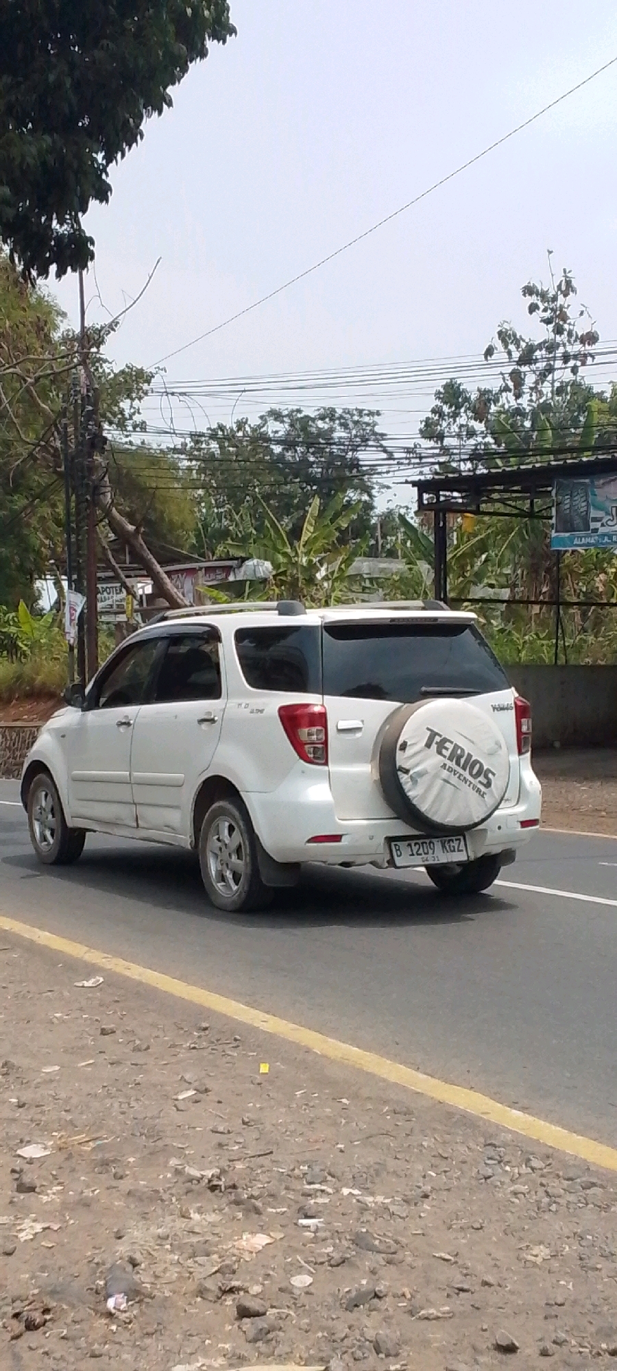 A white Daihatsu Terios with a spare tire cover on the back is parked on the side of a road. The license plate reads "B 1209 KGZ". The car is facing away from the viewer, with the rear of the vehicle clearly visible. The road is paved, with a yellow line marking the edge. The surrounding environment is a mix of vegetation and utility poles. There are trees and bushes on the left and right sides of the road, with power lines visible overhead. In the background, there appears to be a building or a structure with signage. The sky is overcast. The overall impression is a roadside scene in a semi-urban or rural area. It is possible that this image was taken in Cilaku, Indonesia, given the license plate format.