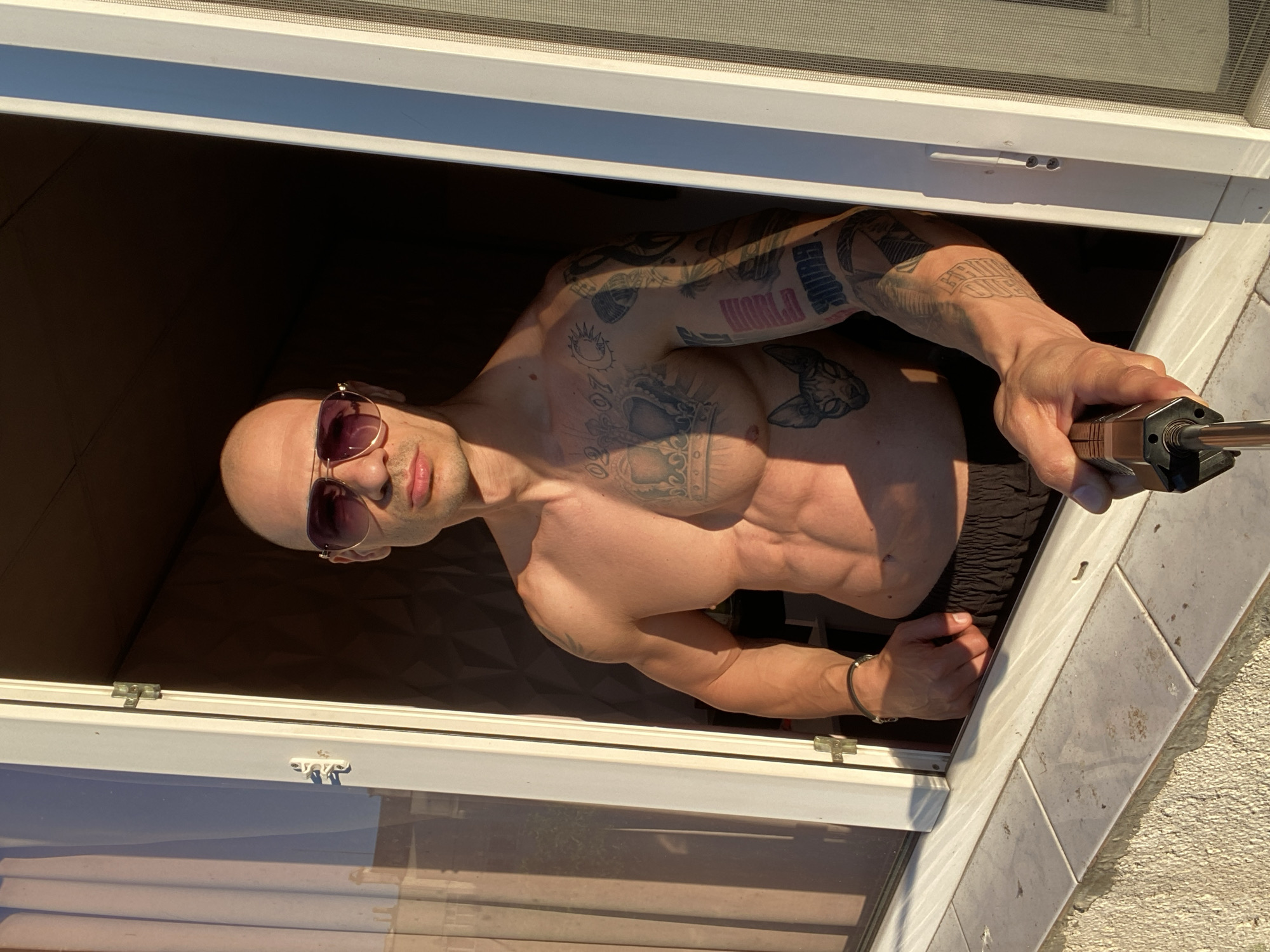 A shirtless, bald man with sunglasses is leaning out of a window. He is muscular and has tattoos on his chest and arms. He is holding a black object in his right hand. The window frame is white, and there is a screen in the top section. The location is unknown and cannot be ascertained.