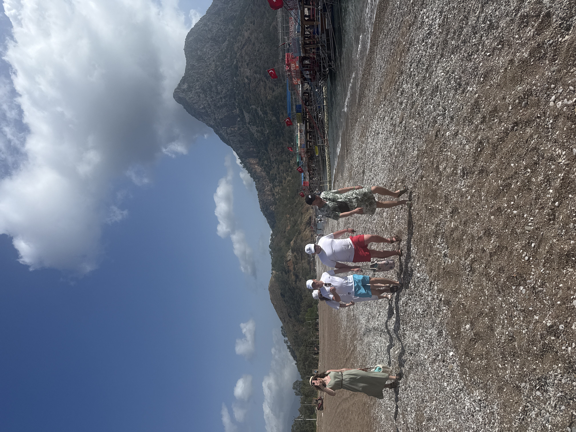 The image depicts a group of people on a beach in Kumluca, Turkey. In the background is a mountain and the sea with several boats anchored along the coast and Turkish flags. The beach is composed of small pebbles and sand. There are five people visible: one person wearing a floral shirt and dark hat, three people wearing white shirts and caps with red or blue shorts and one person wearing a long green dress, facing toward the camera. The sky is blue with white clouds.