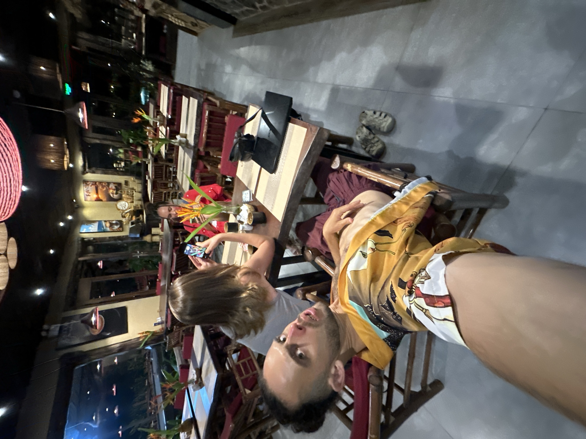 In the dimly lit interior of a restaurant in Nam Hoa Lư Ward, Vietnam, a man in the foreground leans back in a wooden chair, wearing a vibrant yellow and white patterned robe. His eyes are wide open, and his mouth is slightly ajar, as if in mid-sentence or surprise. He is positioned in the lower left of the frame, with his left arm extended towards the camera.
Behind him, a woman with long blonde hair sits at a table, looking down at her smartphone, which displays a colorful image. She wears a grey tank top. Further in the background, another woman in a red top is also seated at a table, appearing to interact with her phone.
The restaurant features numerous wooden tables and chairs, set with placemats and small vases containing orange and yellow flowers. Hanging lamps with woven shades illuminate the space. Large windows offer a view of an outdoor area with more seating and vegetation, likely a garden or patio. The floor is tiled in a light grey. A black laptop is open on a table in the mid-ground, and a black bag rests on a chair. A pair of grey sandals are placed on the floor near the man. The overall atmosphere suggests a casual dining setting in the evening.