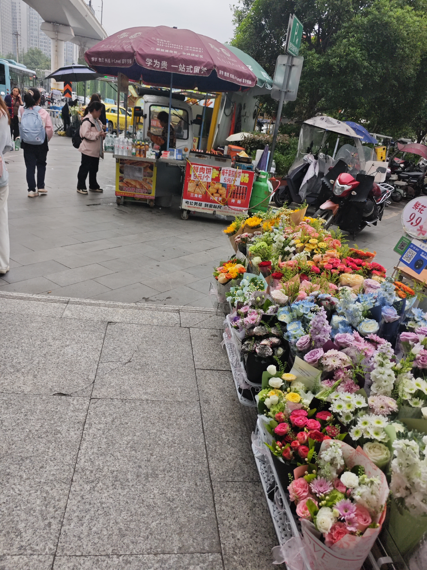 The image shows a street scene in the Chongqing High-tech Zone, China. A flower stand displays various colorful bouquets in white wire baskets. Next to the flowers are several motorbikes parked. In the background, there is a food stall under a red and white umbrella, with people standing around it. A green parking sign is visible. A pedestrian walkway is visible to the left, with a gray tiled surface and a person wearing a pink jacket and a blue backpack, as well as a person wearing white pants. An elevated structure is present above the scene. The stall sells meat pies and sausages. Text on signs includes pricing information for the goods on sale at the food stall and flower stall.