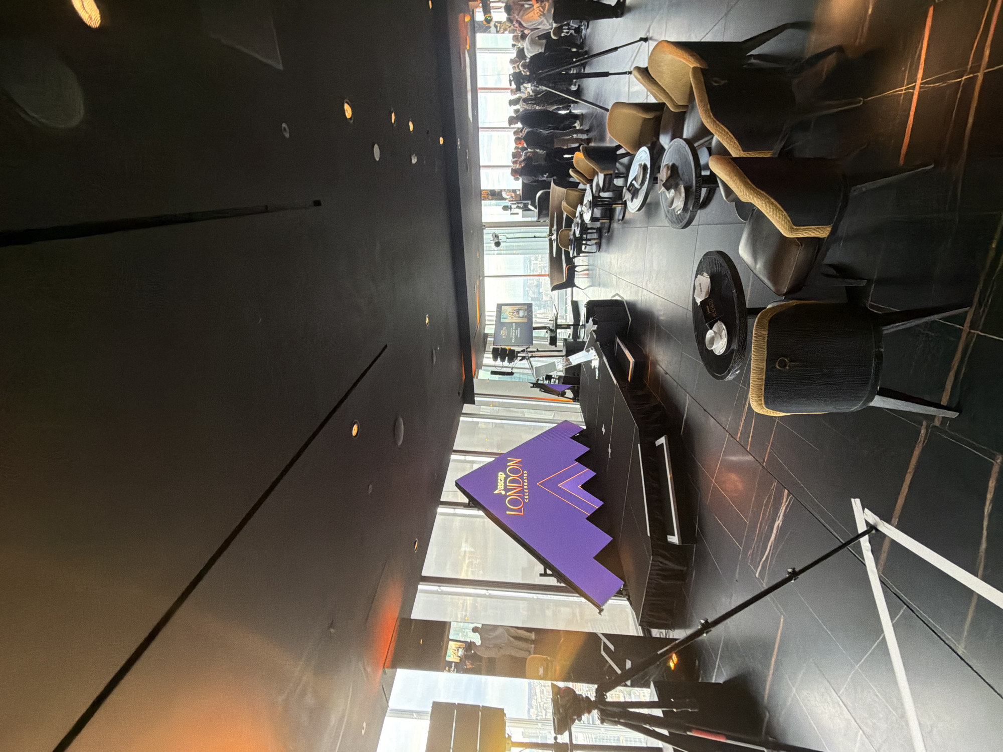The image depicts an indoor event space, likely a venue in London, United Kingdom, situated at a high elevation, as suggested by the panoramic city view visible through large windows. The room features a stage area with a prominent purple triangular backdrop displaying the text "ASCAP LONDON CELEBRATES" in a stylized font. In front of the stage, several round tables with seating arrangements are set up, some with cups and saucers. A diverse group of people are gathered throughout the space, some standing and conversing, others seated. The decor appears modern and sophisticated, with dark flooring and dark-colored furniture. Lighting fixtures are visible on the ceiling, and a camera on a tripod is positioned in the foreground, indicating a live event or recording. The overall atmosphere suggests a formal or semi-formal gathering, possibly a celebratory occasion or a corporate event.