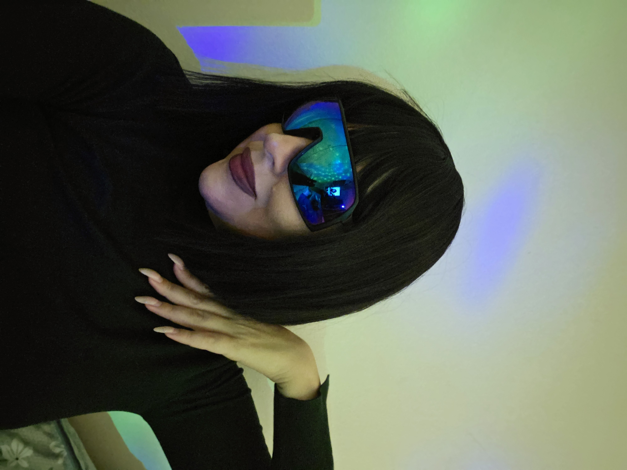 An image, rotated counter-clockwise, depicts a person, likely a woman, with long dark hair and dark clothing (possibly a turtleneck or long-sleeved top). She is wearing prominent, futuristic-looking sunglasses with iridescent, mirrored lenses that reflect blue, green, and purple light patterns. Her face, including her nose and lips adorned with dark lipstick, is visible beneath the glasses. Her right hand, featuring long, light-colored manicured nails, rests near her shoulder or chest area. The background is a wall illuminated by colorful, diffused lighting, primarily in shades of green, blue, and purple, suggesting an indoor setting with mood lighting or a light projection. There is no visible text in the image. The person appears to be posing for the camera.