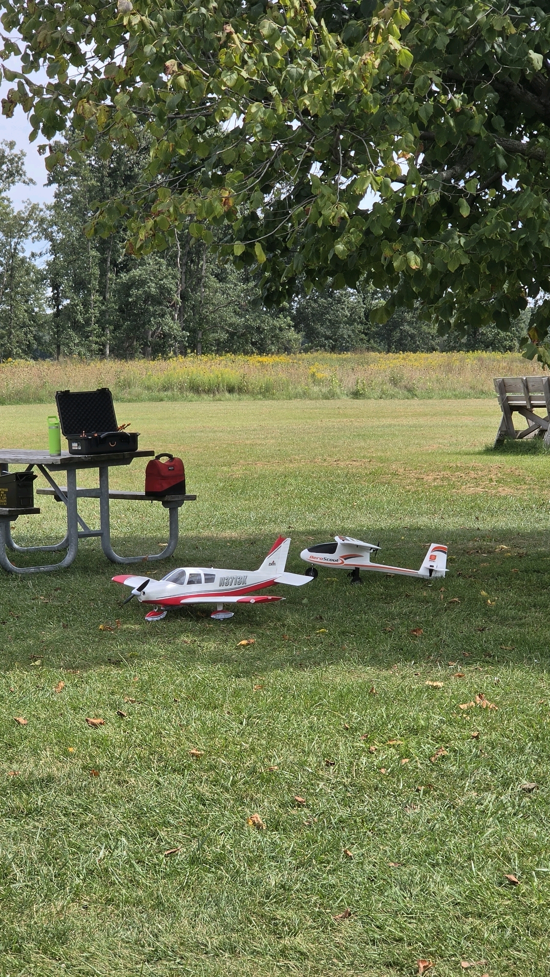 The image depicts an outdoor scene with two model airplanes on a grassy field. A picnic table with a black case, a green water bottle, and a red bag is visible on the left side of the image. The background features trees and a meadow with yellow flowers. A wooden bench is also visible in the distance. The scene appears to be a park in Rolling Meadows, USA, likely set up for a hobbyist activity.
