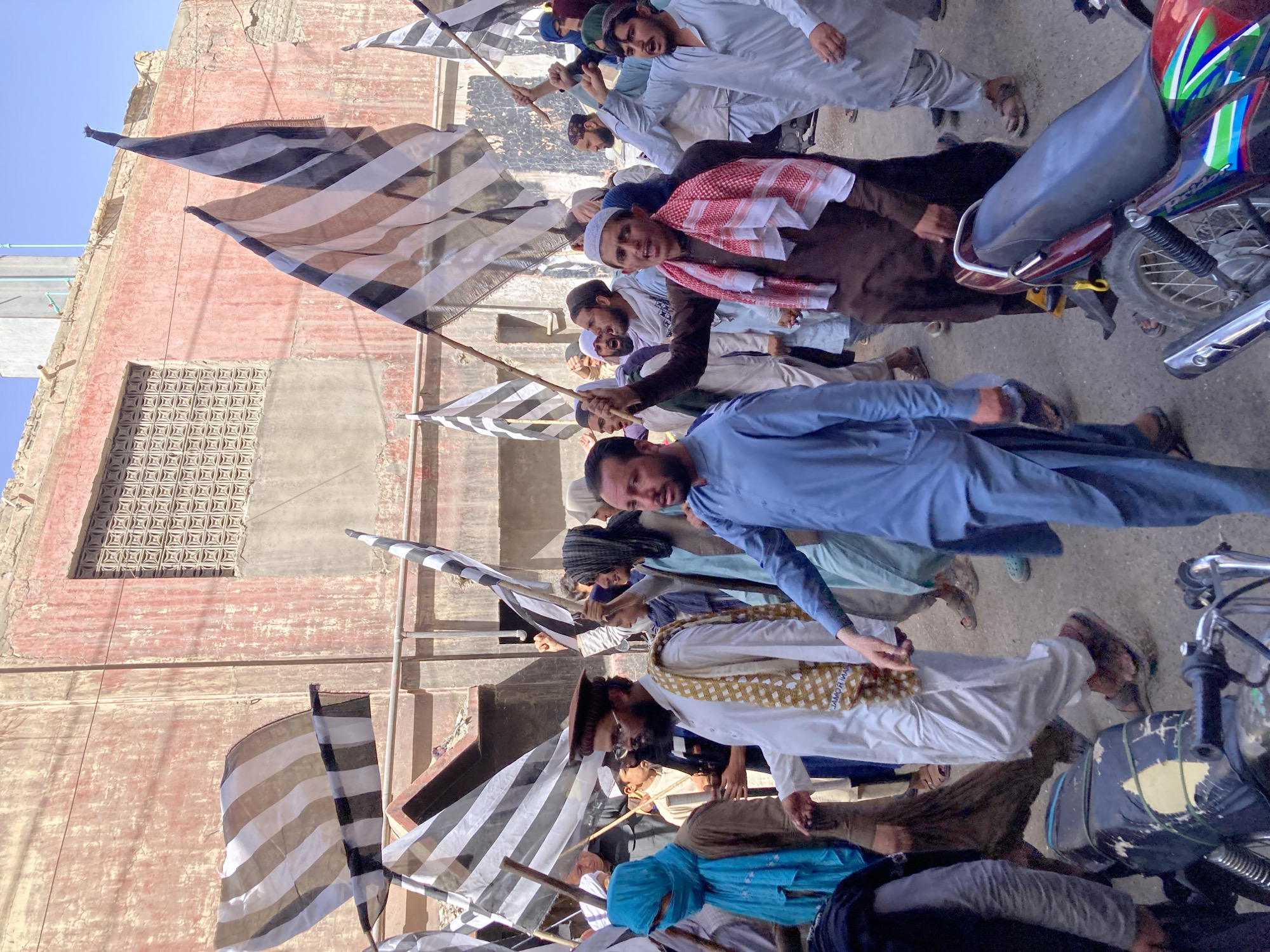 A group of men are marching down a street in Quetta City Tehsil, Pakistan. They are carrying striped flags on wooden poles. The men are wearing a variety of traditional clothing, including robes, skullcaps, and headscarves. Some of the men are smiling and looking at the camera. The street is paved and there are motorcycles parked on the side. A weathered brick building is in the background. The sky is blue.