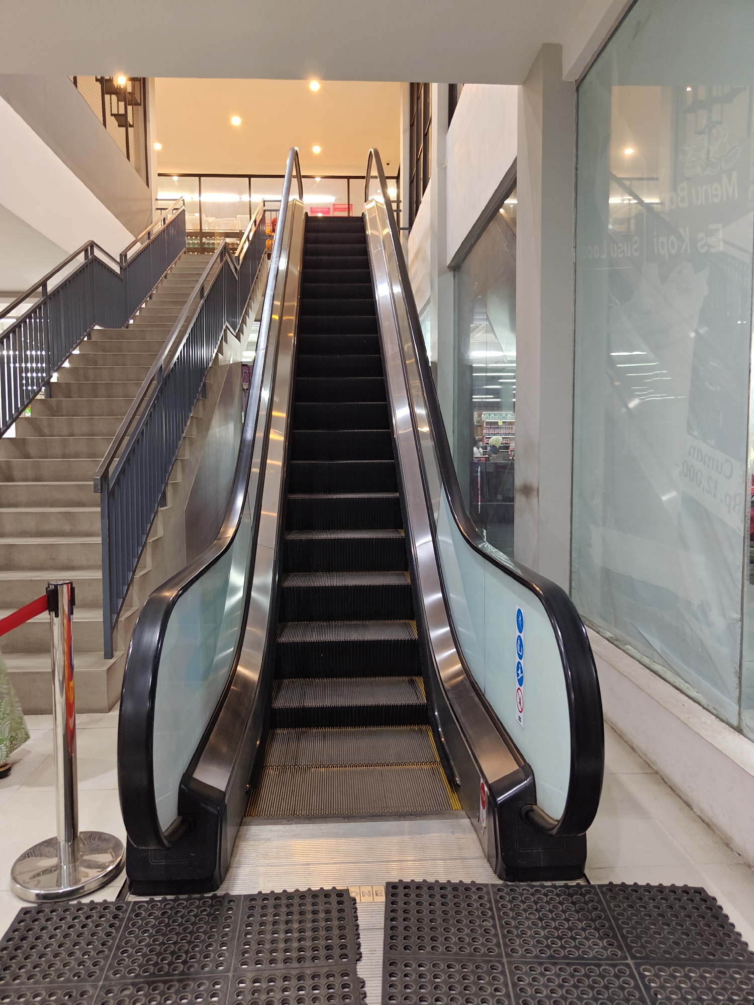 The image captures an indoor scene featuring an escalator and an adjacent staircase, likely within a commercial building such as a shopping mall or a department store, located in the city of Situbondo, Indonesia. The escalator, with black steps and a silver handrail, is in the foreground, angled slightly to the right. To its left, a concrete staircase with a dark grey metal railing ascends. The environment appears well-lit with overhead lights visible. A red velvet rope barrier with a chrome stanchion is positioned to the left of the staircase, suggesting a controlled access area or a queuing point. On the right side of the escalator, a large glass window is partially obscured by a white translucent film bearing text and graphics, reflecting the interior. The floor in front of the escalator and stairs is covered with dark textured mats. In the background, through the escalator and staircase, merchandise shelves and store lighting are faintly visible, indicating a retail setting. No people are prominently featured or engaged in any discernible interaction, though a person's hand and arm, wearing a patterned fabric, are partially visible near the red barrier. The overall impression is of a functional, modern interior space designed for vertical transportation.