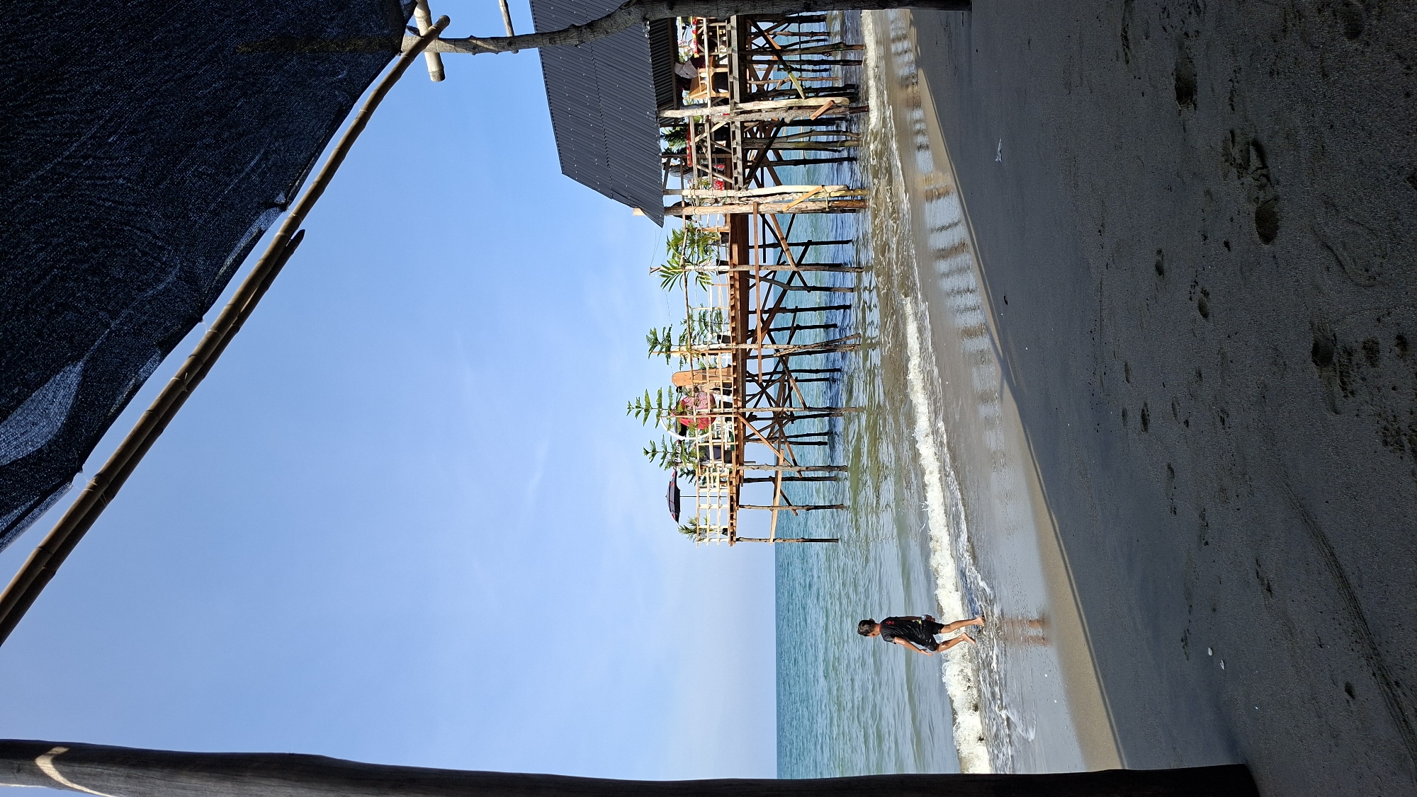 A boy walks on a sandy beach near a wooden stilted structure extending into the ocean. The structure has multiple levels and is adorned with potted plants and some seating areas. A dark canopy stretches across the top foreground of the image, casting a shadow on the beach. The water is calm with small waves lapping at the shore. The sky is clear and blue. The location is a beach in Lhokseumawe, Indonesia.