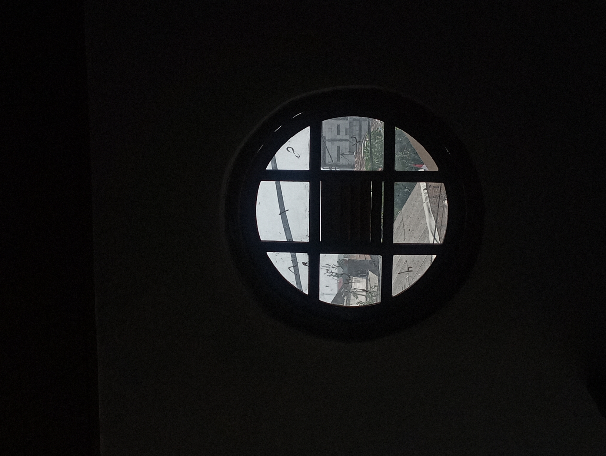 The image is a close-up shot of a circular window with a wooden frame, set against a dark wall. The window is divided into six panes by dark mullions. Through the window, a view of buildings and greenery is visible, suggesting an urban or semi-urban environment. The lighting suggests it is daytime, possibly overcast, given the muted light filtering through the window. There are no people or discernible animals in the image. The style of the window and the architectural elements visible outside hint at a location with some history, possibly in a city like Surakarta, Indonesia, known for its traditional and colonial architecture. The overall mood is somber and contemplative due to the darkness of the surroundings and the filtered light.