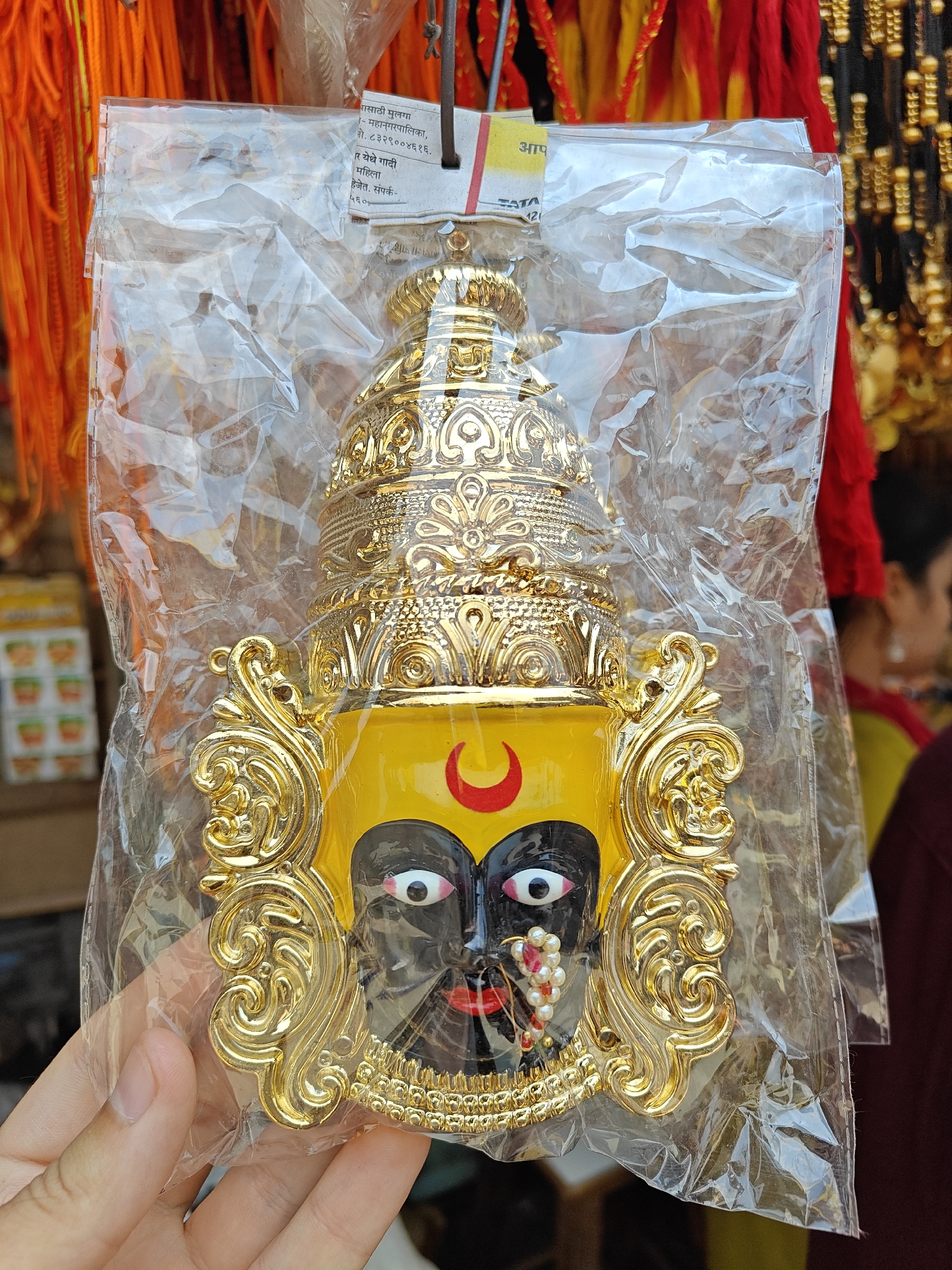 A person's left hand holds a transparent, creased plastic bag containing a decorative, golden-colored plaque depicting an ornate face. The face is black with prominent white eyes, black pupils, and red outlines. A yellow forehead area displays a red crescent symbol. The nose is adorned with a nose ring comprised of white pearls and red beads, and the lips are red. The face is framed by intricate golden scrollwork and topped with a multi-tiered, elaborate golden crown. The item appears to be constructed from molded material with a metallic gold finish and painted details. A white paper tag with red stripes, bearing partially legible text in an Indian script, including "TATA," is affixed to the top of the plastic bag. The background is out of focus, revealing elements of a retail environment, such as hanging orange and yellow decorative cords on the left, and red and yellow fabric elements alongside strands of black and gold beaded items on the right. A blurred, partial figure of an individual wearing red and yellow clothing is visible on the far right. The scene is located in an unspecified city and country.