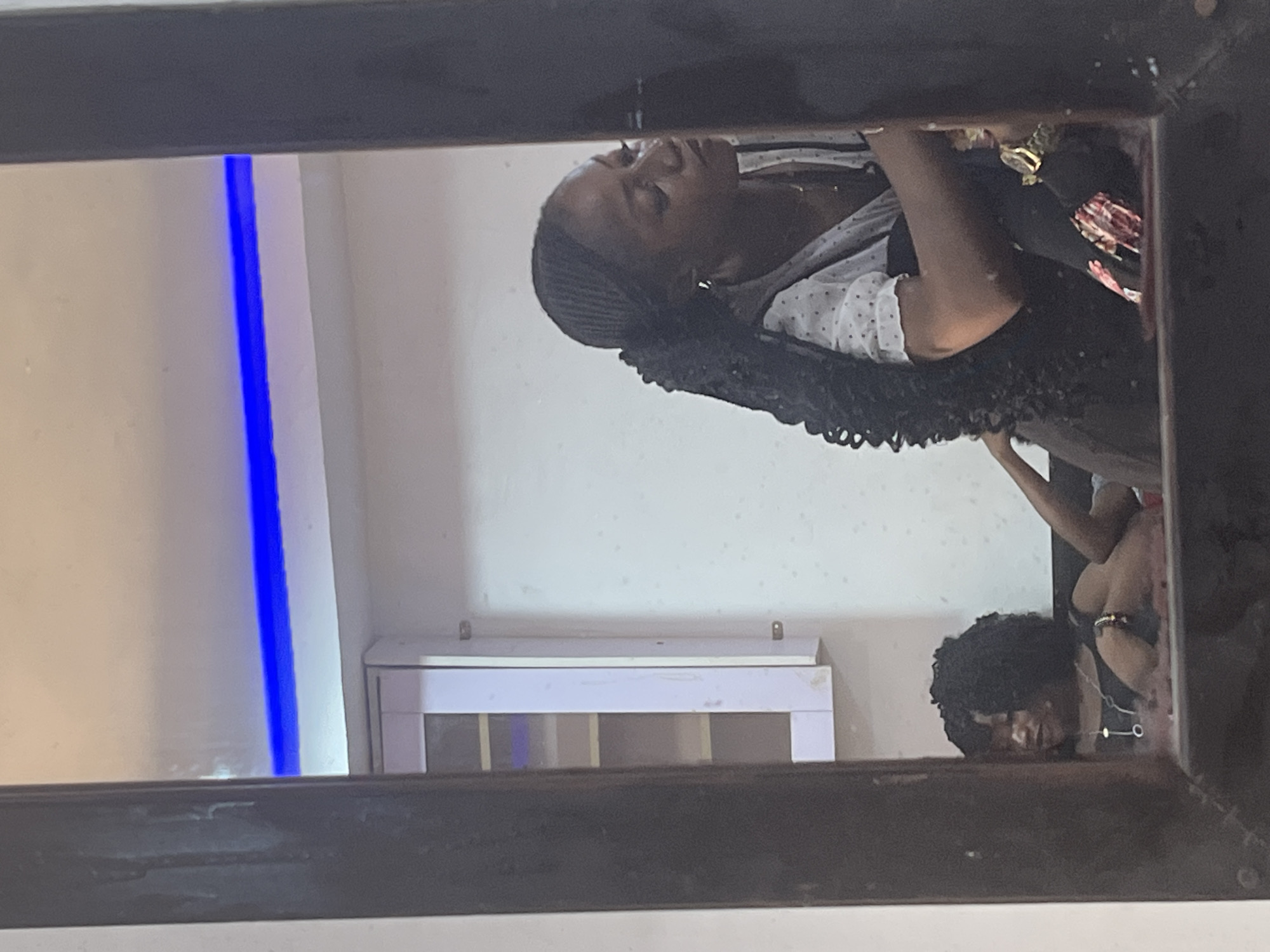 This image captures a reflection in a mirror, set indoors in what is understood to be a location within Abuja, Nigeria. The dark frame of the mirror borders the scene on the left, right, and bottom.
The central subject, a dark-skinned woman, is prominently reflected. She has long, dark, braided hair styled into a high ponytail. Her attire consists of a white, polka-dotted blouse beneath a dark, sleeveless outer garment. A gold-colored watch is visible on her right wrist, along with earrings and a necklace. Her head is turned towards the right, and her gaze is directed outside the visible reflection, indicating a focused attention.
In the lower-left portion of the reflection, a second dark-skinned woman is visible, seated or reclining. She has dark, curly hair and wears a dark top, a necklace with a circular pendant, and a bracelet on her left wrist. A bare leg is partially visible.
The reflected background features a light-colored wall. A horizontal blue LED strip light illuminates the upper section of the wall or ceiling. To the left of the main subject's reflection, a white, multi-compartment wall-mounted cabinet is visible, with a purple item discernible within one compartment. A small piece of pink or red material is visible at the bottom center of the reflection, near the main subject's arm. The interior space appears adequately lit.