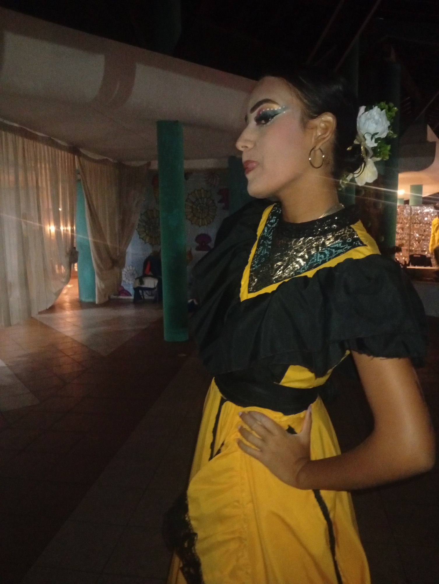 A young woman in a traditional yellow and black dress stands in a dimly lit indoor space. The dress features a sequined collar, puffed sleeves, and a sash at the waist. She wears gold hoop earrings and a white flower in her hair. Her makeup includes dramatic eyeshadow and lipstick. She is in a profile view, looking to the left, with her hand resting on her hip. The background shows tiled flooring, sheer curtains, and turquoise support columns. A decorated wall with floral designs is partially visible. It's dark, suggesting evening or nighttime. This image was most likely taken in Playa El Agua, Venezuela.