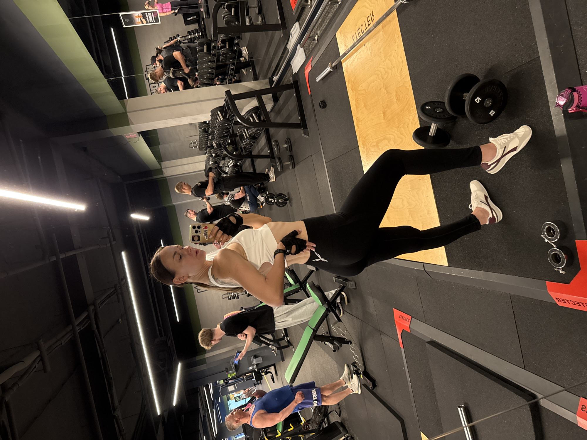 This is a full-body selfie taken in a gym. The photographer, a woman in athletic wear, is standing and holding a smartphone in her right hand. She is wearing a white tank top, black leggings, and white and maroon athletic shoes. She has black fingerless gloves on. The gym appears to be well-equipped with various weight machines, dumbbells, and a bench press. In the background, other gym-goers can be seen exercising. The environment is brightly lit with overhead fluorescent lights and some natural light entering from large windows, which are not visible in the frame. The floor is covered with black rubber mats. The gym appears to be modern and well-maintained. There is no information to suggest this is located in Sochi, Russia.