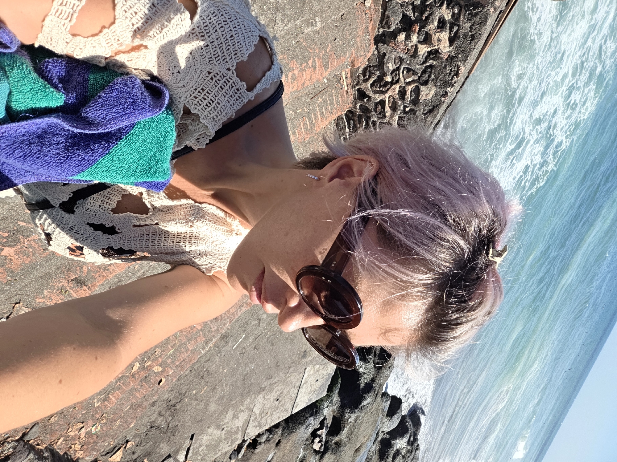 A woman with short, pink-tinted hair and large, dark sunglasses is seated on concrete steps near the ocean. She is wearing a white, crocheted top and holding a folded towel with purple and green stripes. The ocean, with its choppy turquoise waves, is visible in the background. Rocky terrain and more concrete structures are also present in the environment. The location is identified as North Kuta, Indonesia, likely near a coastal area.