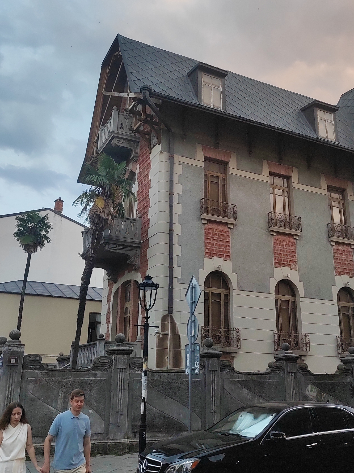 The image captures a street view in Batumi, Georgia, on an overcast day. Dominating the frame is a multi-story building with a dark, patterned roof and dormer windows. The facade of the building is a mix of grey stucco and red brick accents, with arched windows on the lower level and rectangular windows with small decorative balconies on the upper levels. A prominent balcony juts out from the upper corner of the building, adorned with intricate stonework. Two palm trees are visible, one tall and slender, and another shorter, fuller one, leaning against the building.
In the foreground, a man and a woman are walking hand-in-hand on a sidewalk. The woman is wearing a white sleeveless dress, and the man is in a light blue polo shirt and light-colored pants. They are walking away from the camera, towards the left side of the image. To their right, a black car is parked at the curb. The street is lined with an ornate stone fence with decorative pillars. A traditional-style lamppost stands next to the fence. The sky is cloudy, contributing to a subdued lighting condition. The overall impression is that of an older European city with a touch of tropical influence.