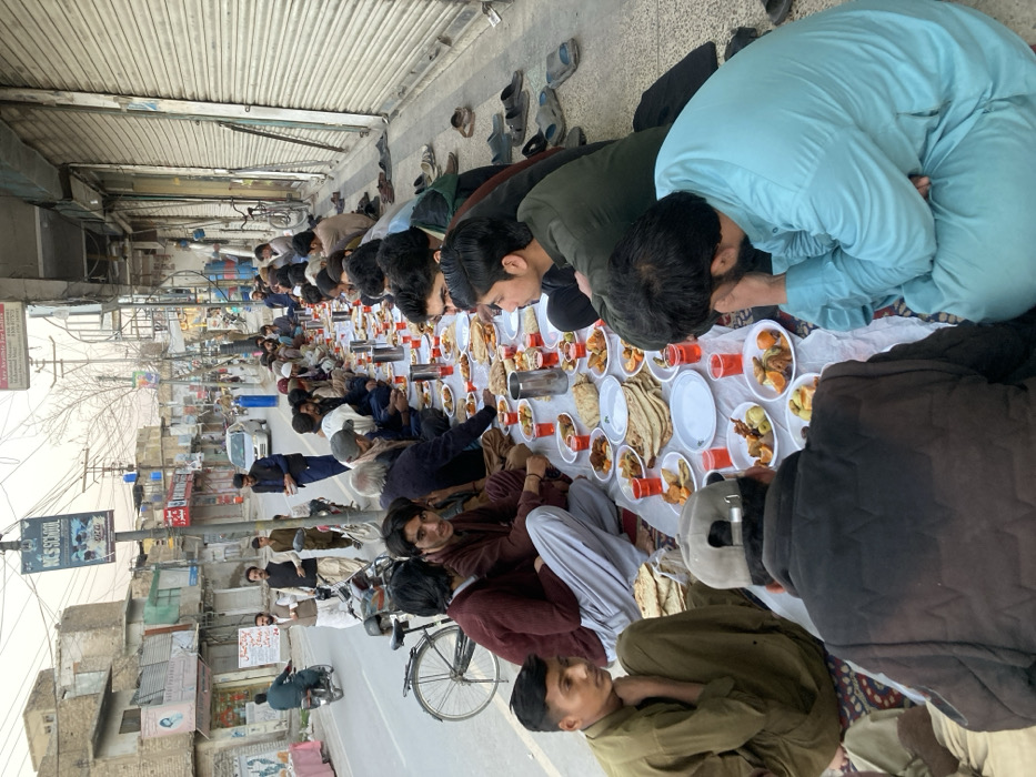 The image depicts an outdoor communal meal occurring on a sidewalk adjacent to a street in Quetta City Tehsil, Pakistan. Approximately 30-40 adult males are seated cross-legged on patterned rugs or mats, arranged in two long rows facing each other. A long white sheet, serving as a tablecloth, is spread over the rugs. Numerous white plates filled with food are distributed along the cloth, including flatbreads (naan), various cooked dishes containing meat and vegetables, and cut fruit (oranges, apples). Small clear plastic cups containing a red beverage and several metallic jugs are also present on the table. Individuals have removed their footwear, consisting of sandals and shoes, which are placed behind them on the sidewalk.
In the background, a paved street runs parallel to the dining area. A white car and a motorcycle are visible on the road, with another bicycle parked near the diners. Several individuals are standing on the street and sidewalk. The street is lined with multi-story buildings; some appear residential, while others are commercial establishments, many with their metal roller shutters closed. Overhead, utility poles and a dense network of power lines are visible. A blue and white sign for "NCS School" is mounted on a pole above the street. The sky appears overcast or hazy, indicating either late afternoon or early evening light conditions.