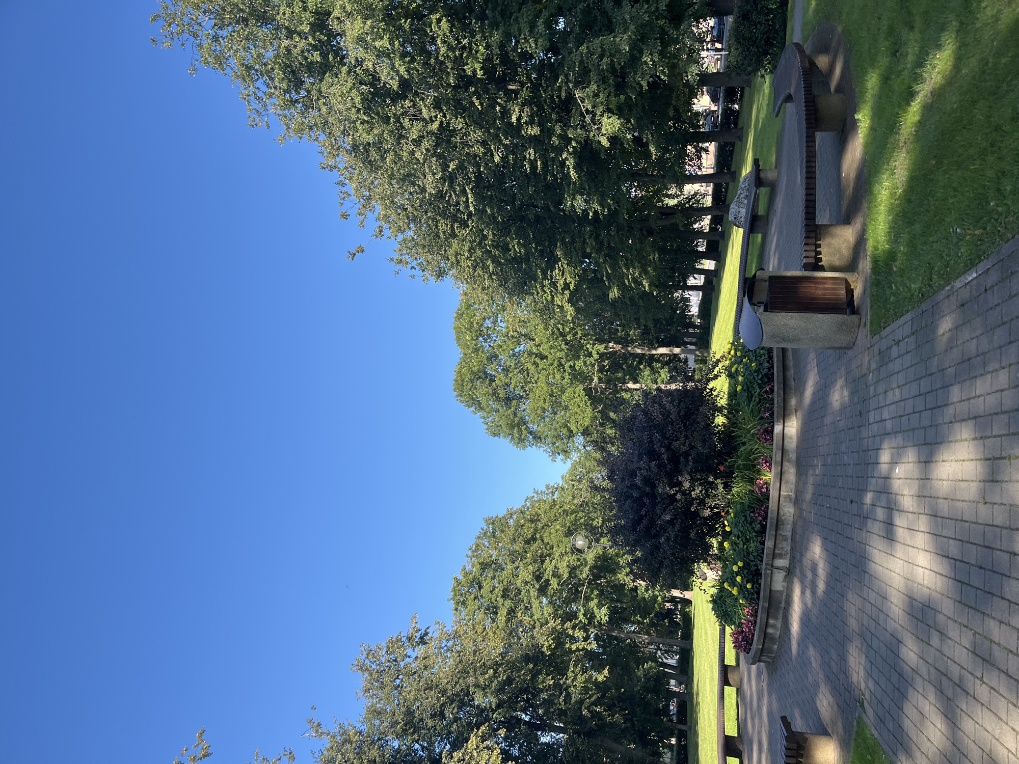 This is a photograph of a public park in Kohtla-Järve linn, Estonia, taken on a sunny day. The park features a paved walkway with a circular flower bed in the center, adorned with yellow and pink flowers and a dark-leaved shrub. Surrounding the flower bed are curved benches made of wood and stone, with one bench featuring an integrated trash bin. In the background, a line of tall, mature trees creates a canopy against a clear blue sky. The park also includes grassy areas and more trees further into the background, with glimpses of urban elements like cars and buildings suggesting its location within a city. The overall impression is one of a well-maintained, peaceful urban green space.