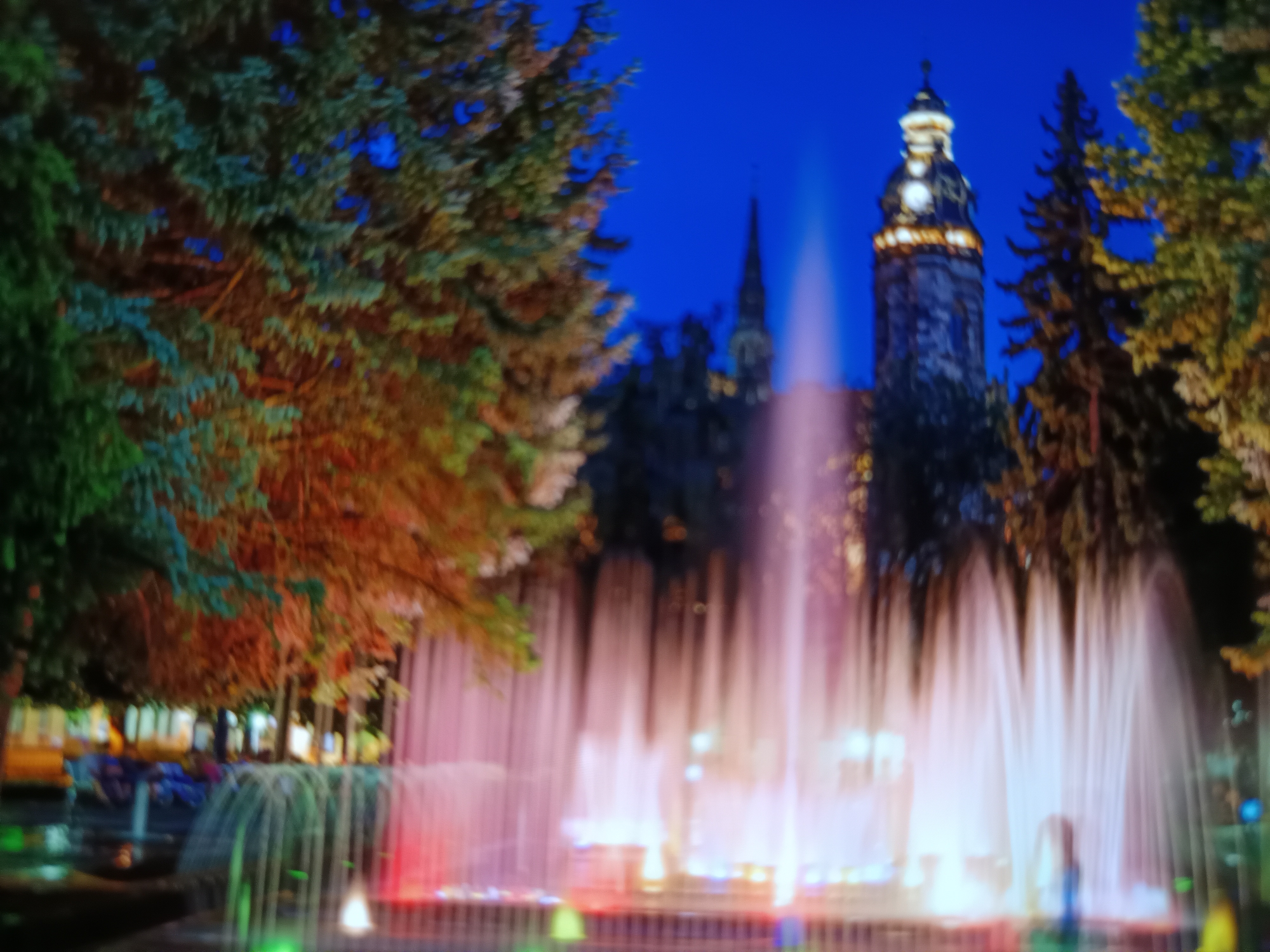 The image shows a night view of a fountain with colorful lights, set against a backdrop of trees and a building with a tower. The fountain's water jets are illuminated with pink and white light, creating a soft glow. In the background, a tall building with a clock tower is visible, its windows lit up. The scene is framed by dark trees, some of which have warm-toned lights reflecting on their foliage. The overall impression is one of a serene and picturesque urban setting at night. This scene is located in the city of Košice, Slovakia, specifically at the fountain in front of the State Theatre of Košice.