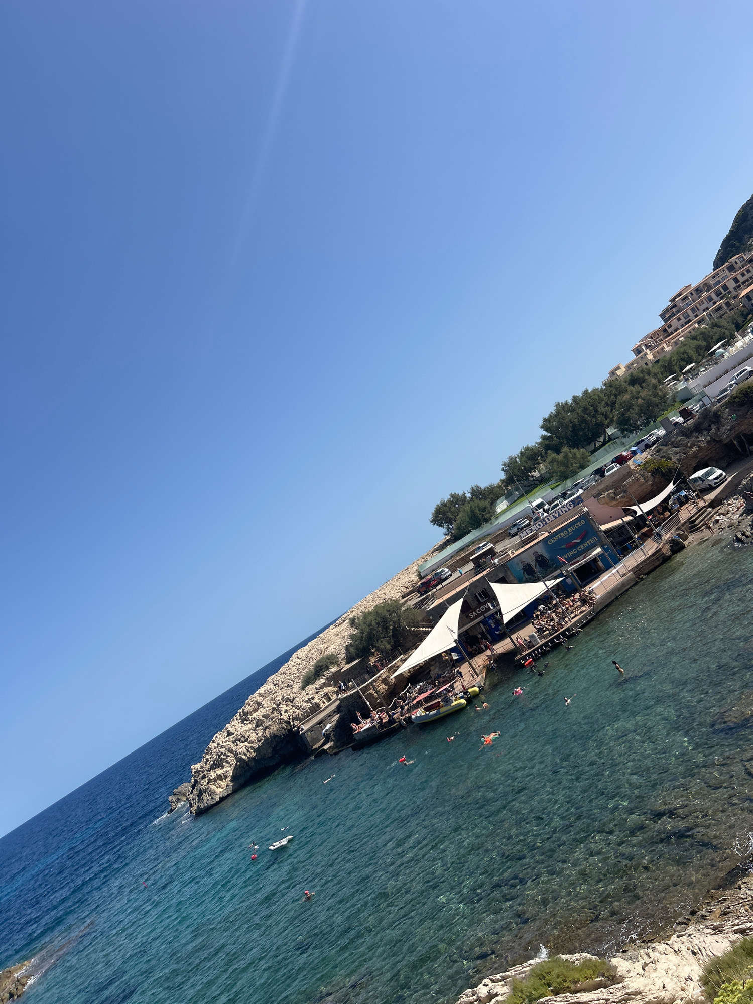 The image depicts a coastal scene in Capdepera, Spain, on a clear, sunny day. The foreground features the turquoise waters of the Mediterranean Sea, with several individuals swimming and floating. A small inflatable raft is also visible in the water. The coastline is rocky, with some vegetation.
Further inland, a cliffside is developed with a multi-level resort or hotel complex, characterized by beige buildings with red-tiled roofs. Below the resort, a row of parked cars is visible on a paved area. Adjacent to the water's edge, a structure with white awnings and outdoor seating areas serves as a beach club or diving center. Signs on this structure read "CENTRO BUCEO" and "DIVING CENTER". People are seated at tables and standing around this area. The overall impression is a vibrant, recreational coastal environment.