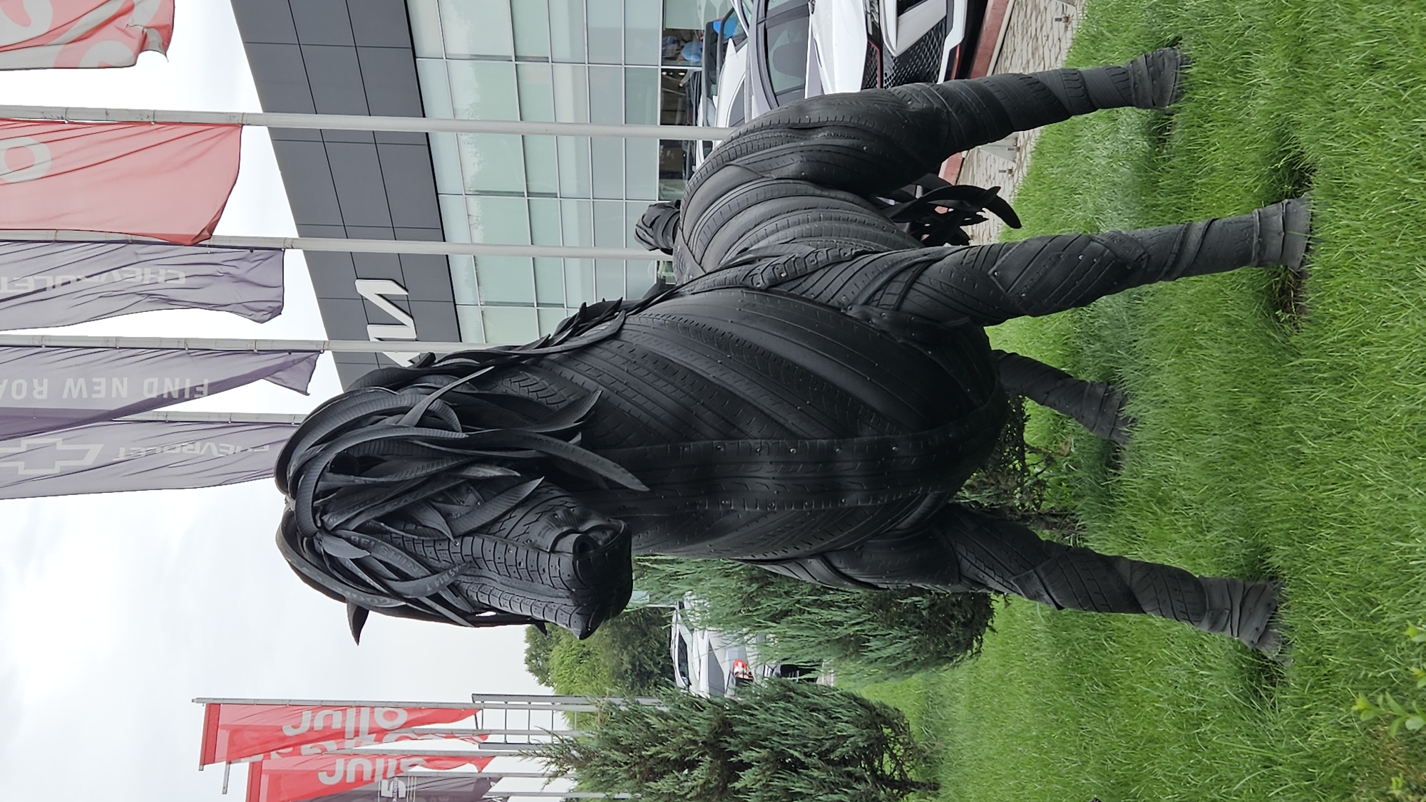 An outdoor sculpture of a horse made from black tires stands on a lush green lawn. The sculpture is detailed, with the mane, body, and legs all constructed from various tire components. Behind the sculpture, a modern building with large windows and the "KIA" logo is visible. Several flags, including red and purple ones with text like "CHEVROLET" and "FIND NEW ROAD," are planted in front of the building. Several cars are parked in front of the building, suggesting a car dealership. The sky is overcast. The location is confirmed as a KIA dealership in Almaty, Kazakhstan. There are no people clearly visible in the image.