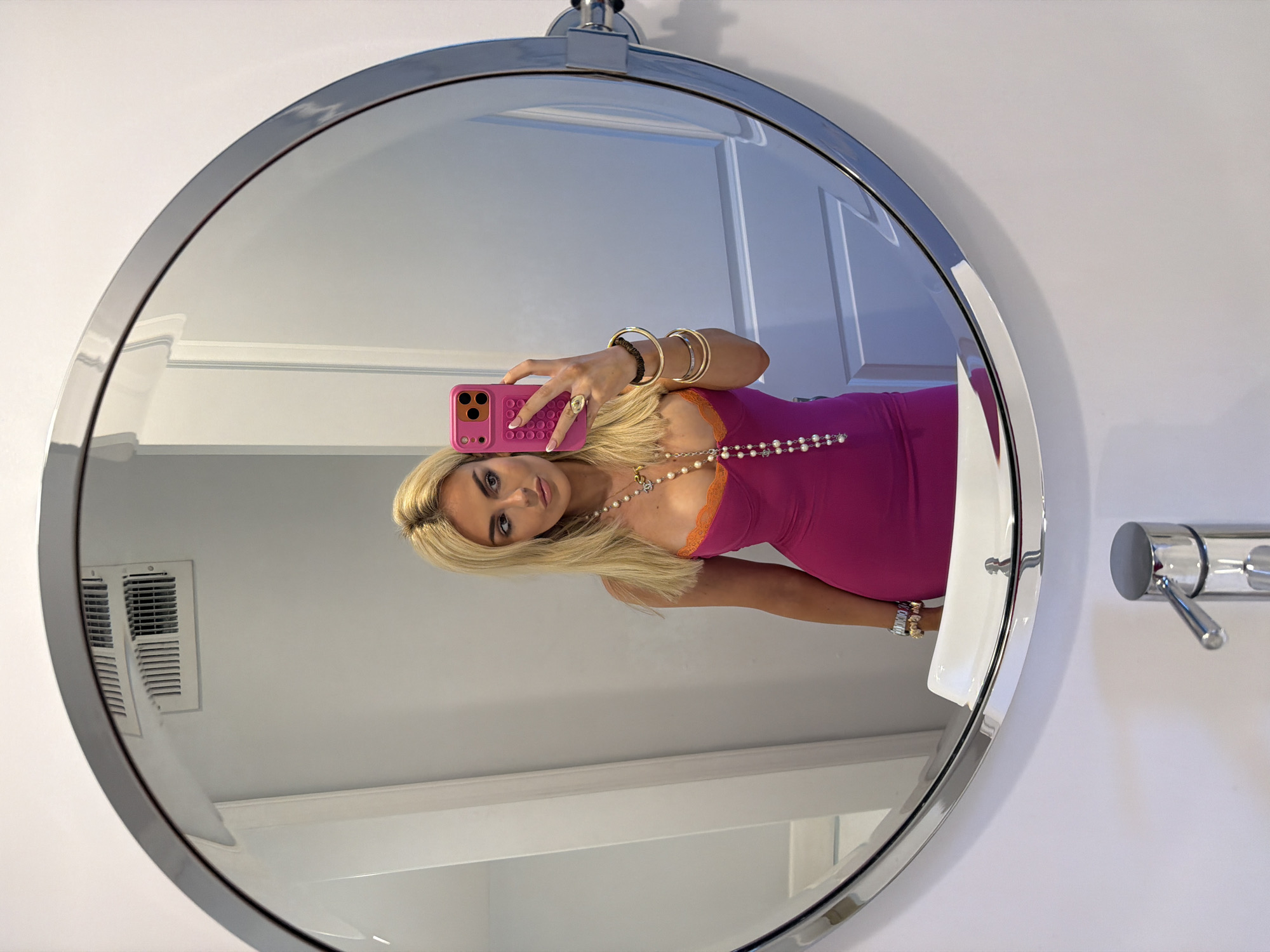 A mirrored selfie taken in a bathroom, likely in a home located in Los Angeles, California. The scene features a woman reflected in a round mirror. She has long blonde hair, wears a pink dress with an orange lace trim, a pearl necklace, multiple bracelets, and large gold bangles. She is holding a pink cell phone, taking the picture. The bathroom has white walls, a white sink, a chrome faucet, a wall vent, and a door with white trim reflected in the mirror.