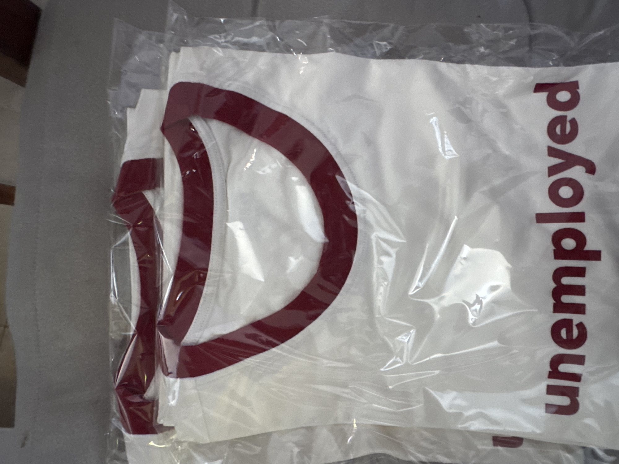A close-up, top-down view of two white t-shirts with burgundy neck and sleeve trim, both packaged in clear plastic. The t-shirts are layered on top of each other, with the bottom one partially visible. The word "unemployed" is printed in burgundy lettering on the front of the bottom t-shirt. The background is a gray, textured surface, likely a bed or floor. There are no people or discernible environmental features indicating a specific location, therefore no location in Kuta Selatan, Indonesia can be determined.