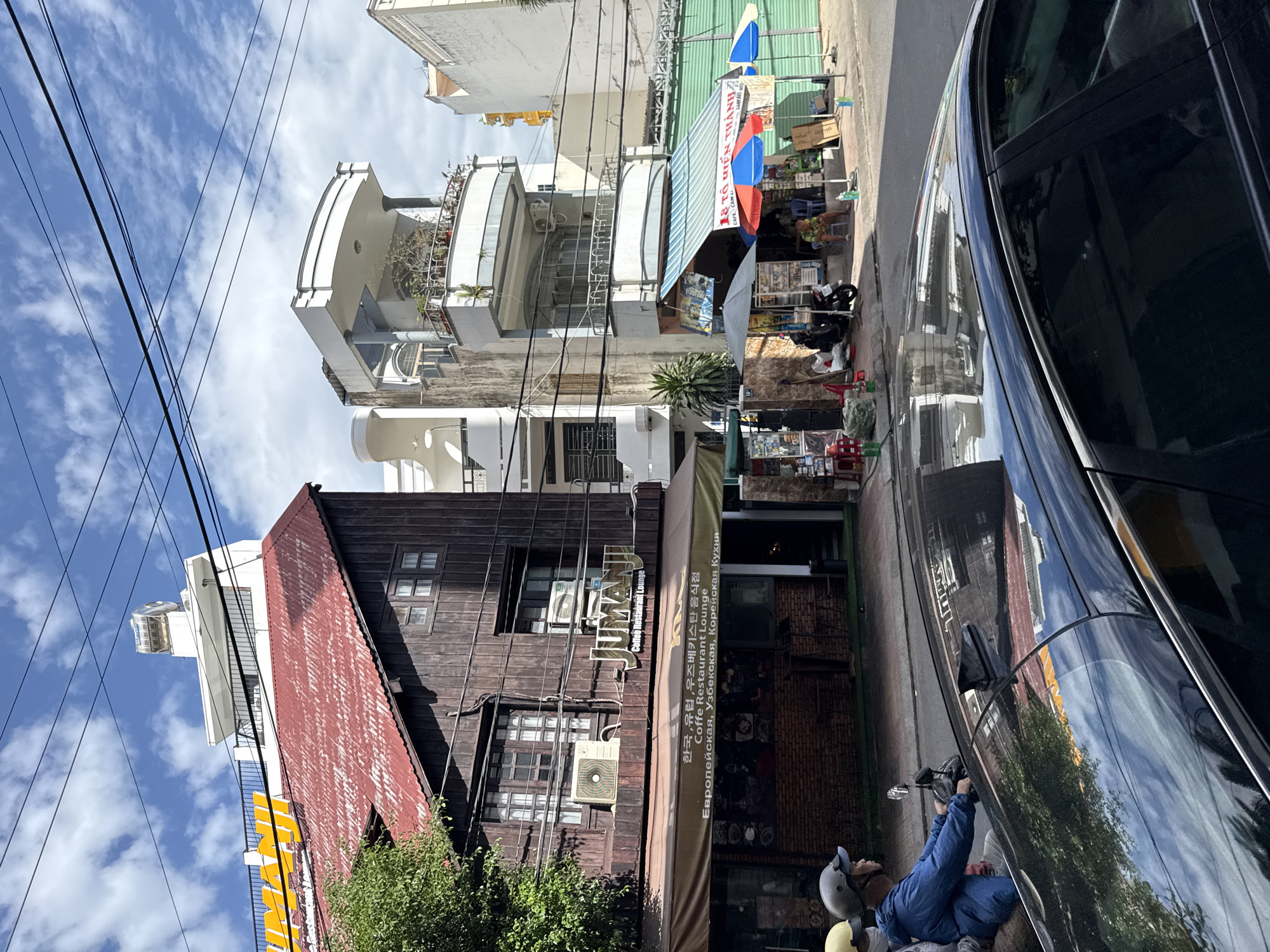The image captures a street scene in Nha Trang, Vietnam, with a focus on the architecture and businesses lining the road. On the left, a dark wooden building with a red corrugated metal roof is prominent, displaying a "JUMANI Coffee Restaurant Lounge" sign. Below this, an awning displays text in Korean, Russian, and English, indicating a European, Uzbek, and Korean cuisine restaurant. Adjacent to this building, a modern white multi-story structure with balconies stands. Further to the right, a smaller shop with striped awnings and colorful umbrellas is visible, appearing to be a cafe or small eatery. A black car occupies the foreground, with reflections of the buildings and sky visible on its polished surface. In the middle ground, a person wearing a blue jacket and helmet is riding a scooter from left to right, their form partially obscured by the car. Several other individuals are seated or standing near the shops on the right side of the street. The sky above is a bright blue with scattered white clouds, and power lines crisscross the upper portion of the image. The overall impression is a bustling urban environment with a mix of traditional and modern architectural styles.