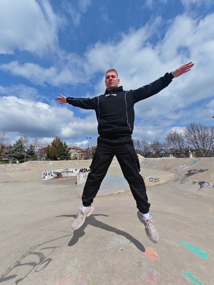 A single adult male is depicted in mid-jump at a concrete skatepark in Shumen, Bulgaria. He is centrally positioned, with both feet elevated off the ground, arms extended horizontally, and legs spread in an 'X' shape. The male wears a black hooded sweatshirt with white double stripes on the sleeves and a white chest logo, black cargo pants, and light-colored athletic shoes. Shadows from his body are cast on the concrete surface. The skatepark consists of worn concrete ramps and flat areas, heavily marked with multi-colored graffiti, including text such as "HOME" and "OMKAHA". In the background, a park area with mostly bare deciduous trees and some evergreen trees is visible, along with residential buildings and streetlights. The scene is brightly illuminated under a blue sky with scattered white cumulus clouds.