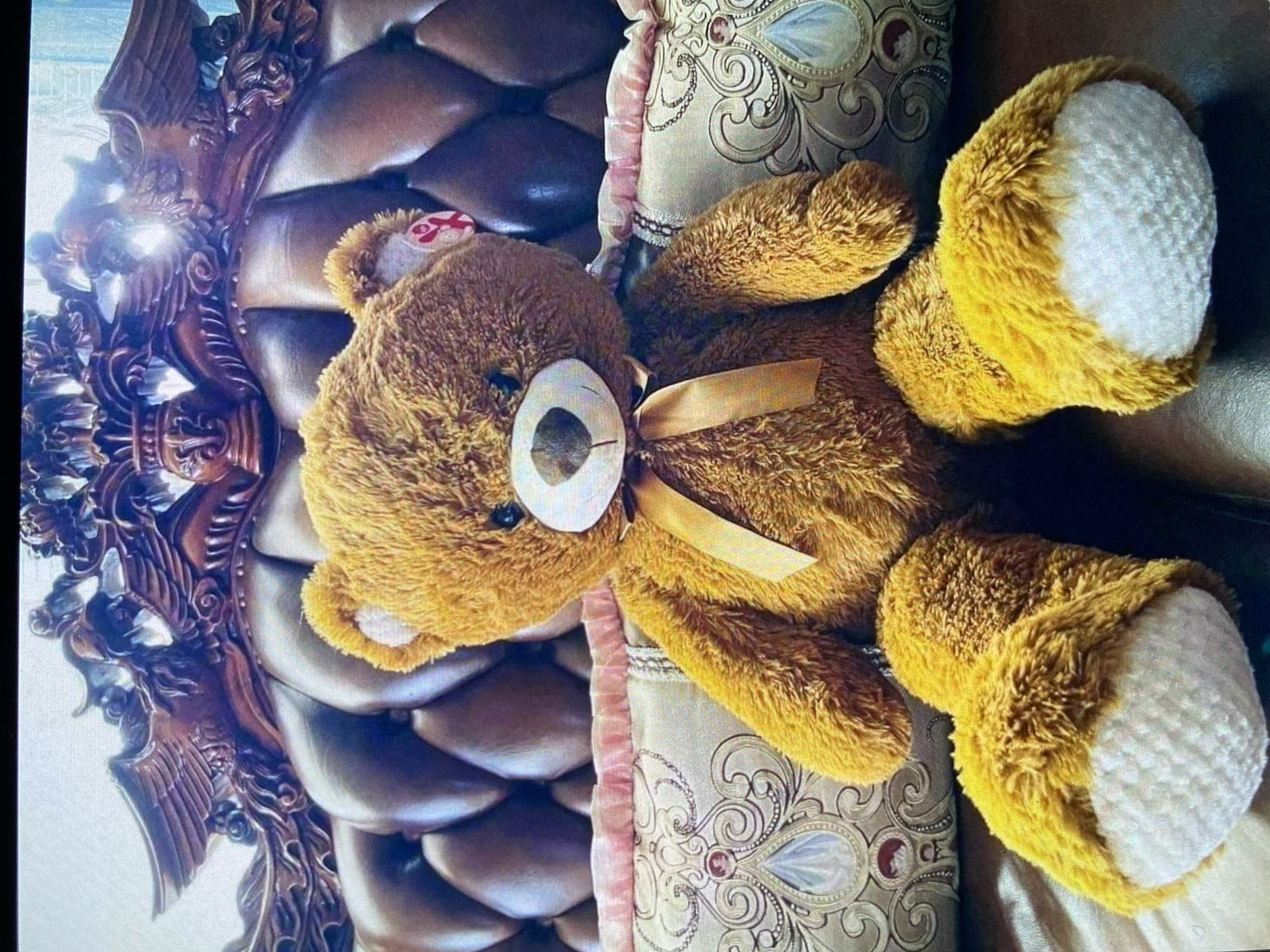 A warm-toned image showcases a fluffy brown teddy bear resting comfortably indoors. The bear is positioned lying down, with its head towards the left side of the frame and its body extending diagonally to the right. It features a light-colored snout with a dark nose and eye, and a golden ribbon tied around its neck. Its paws and feet also have white or cream-colored undersides, providing a contrast to its brown fur.
The teddy bear is placed on what appears to be a soft, textured surface, possibly a bed or a sofa. To the bear's right, a decorative cushion or pillow is visible, adorned with an intricate cream-and-gold patterned fabric and subtle pink piping along its edge. Behind the bear and to its left, an elaborately carved dark wooden structure forms part of the background, suggesting a piece of ornate furniture like a headboard or chair frame, consistent with a traditional interior design. The wood exhibits reflections from what appears to be soft indoor lighting.
No human activity is depicted; the scene is still and focuses solely on the teddy bear and its immediate surroundings. A small red tag with white markings, possibly a brand label, is partially visible on the back of the teddy bear's head, peeking out from its fur. The overall color palette is rich with browns, golds, and creams, creating a cozy and luxurious atmosphere. While the image does not contain specific geographical markers, this indoor scene is contextualized as being in Bankya, Bulgaria.