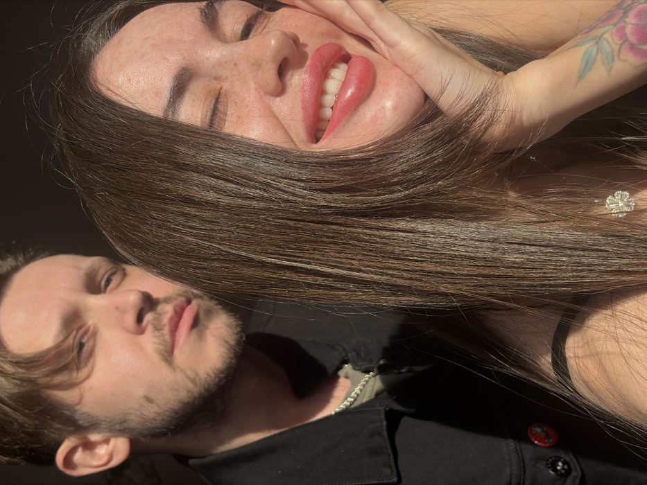 The image captures a close-up indoor portrait of two individuals, a male and a female, reportedly located in the city of Saint Petersburg, Russia. Direct, strong sunlight illuminates the scene from the right, casting highlights and shadows across both subjects.
In the foreground, occupying the right half of the frame, is a female with long, dark brown hair flowing down her right side. Her eyes are closed or squinted due to the bright light, and she is smiling broadly, revealing her upper teeth and some lower teeth. Her left hand is positioned against her chin and jawline. Her skin features visible freckles. She wears a dark, possibly black, strap garment, and a silver-toned, four-leaf clover-shaped pendant is visible around her neck. A tattoo with floral patterns is present on her left forearm.
Behind and to the left of the female, partially obscured by her hair, stands a male. He has light brown hair and a short beard, and he looks directly towards the camera with a neutral expression. He is dressed in a dark jacket, likely denim, worn over a lighter-colored shirt, with a silver chain visible around his neck. A circular red and black pin or badge is attached to the left side of his jacket. The background is uniformly dark and out of focus, indicating a shallow depth of field.