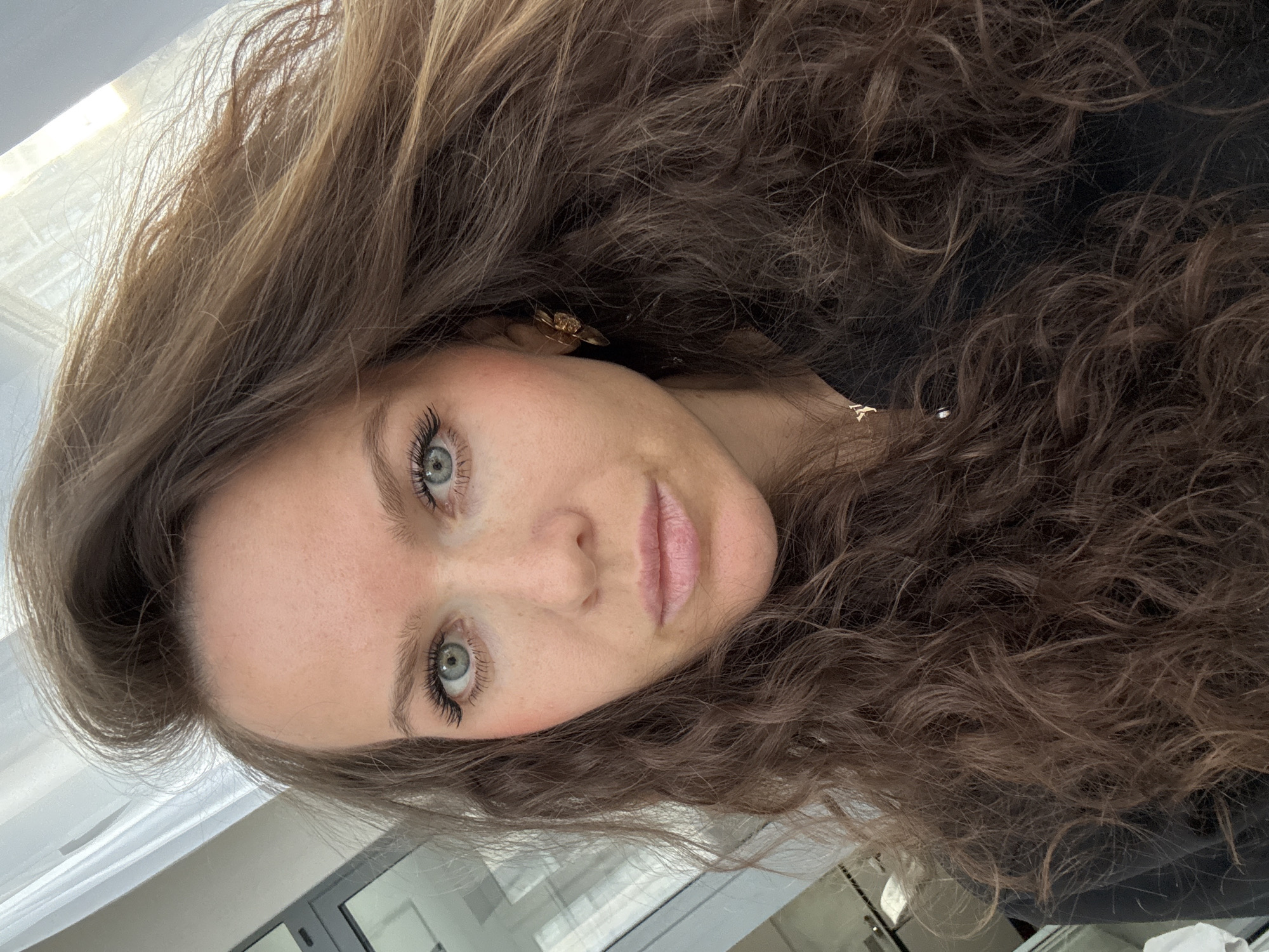 A close-up portrait features a young woman with long, wavy, voluminous brown hair that cascades around her face and shoulders. Her head is tilted, and she gazes directly at the viewer with striking light blue or grey-green eyes, accentuated by prominent dark eyelashes. Her complexion is fair, with a hint of pink blush on her cheeks and light pink lipstick on her lips. A small, golden-colored earring is partially visible on her right ear, nestled within her hair. She wears a dark top, possibly black. The background is softly blurred, suggesting an indoor setting with natural light entering from a window, visible as light reflections and structural cues. Her expression is calm and neutral. No text is visible in the image.