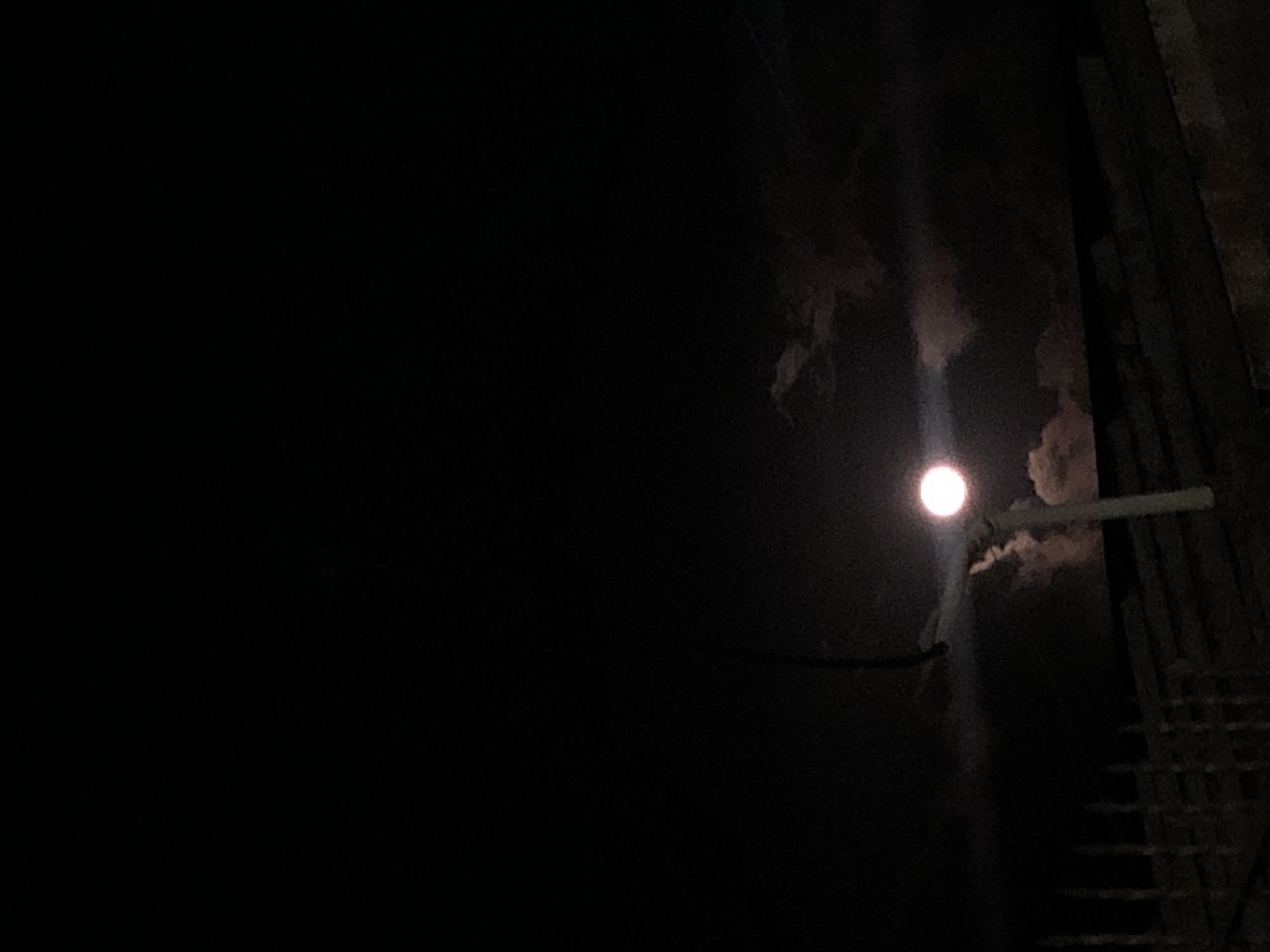 In a dark environment, a round light source is present, positioned near a structure that appears to be made of wood. This structure is composed of vertical and horizontal bars, resembling a ladder or scaffolding. The dark background creates a stark contrast with the light source, which illuminates a small area around it. Faint cloud formations are visible above the light source. A wire extends from left to right across the lower part of the illuminated section. Location details not available.